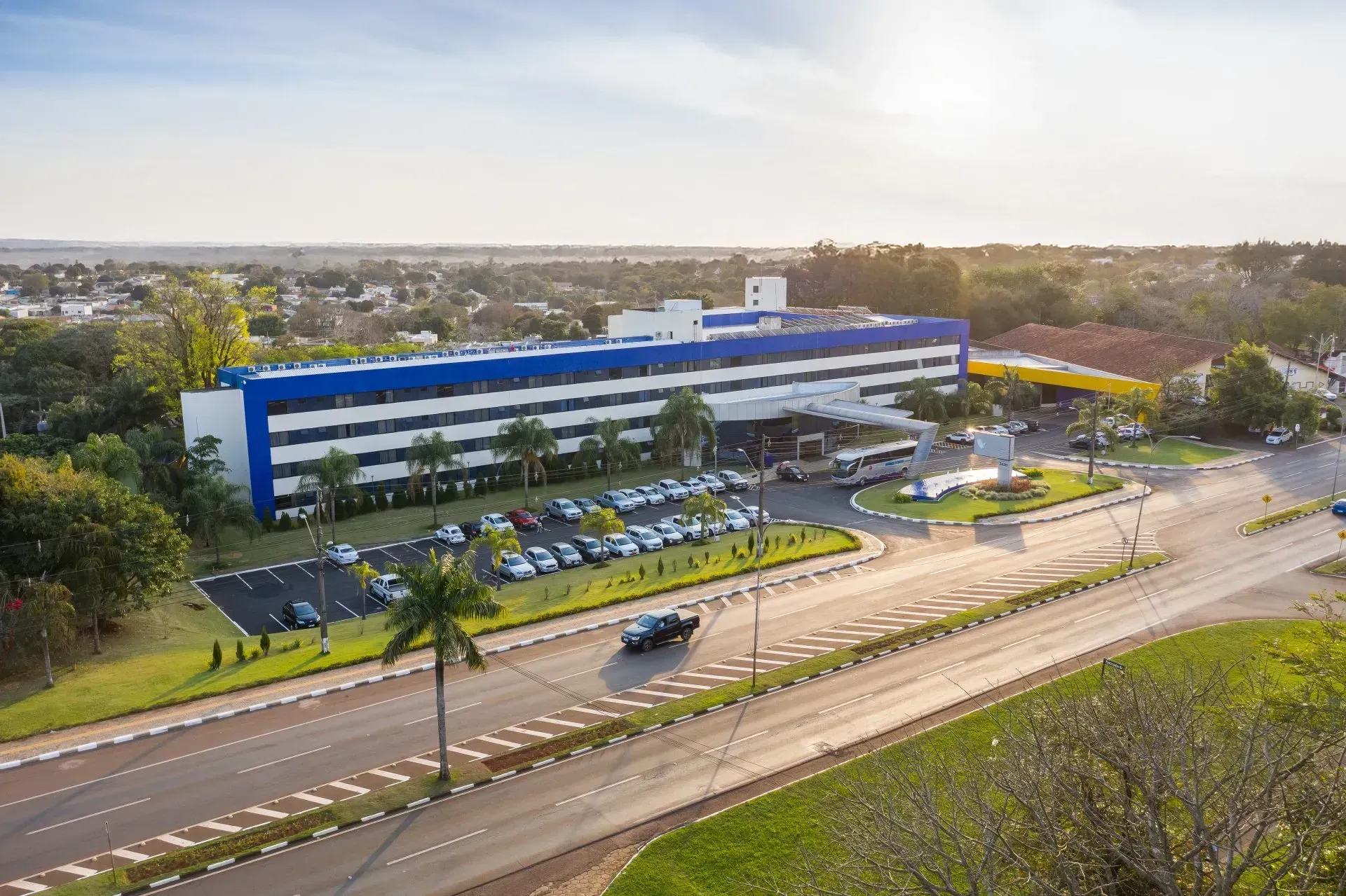 Prédio azul e branco com um amplo estacionamento, palmeiras e uma rua em frente. Dia ensolarado.