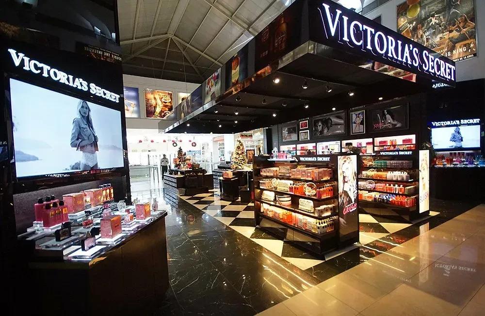 Interior da loja Victoria's Secret. Piso xadrez preto e branco, expositores de produtos de beleza e sinalização.