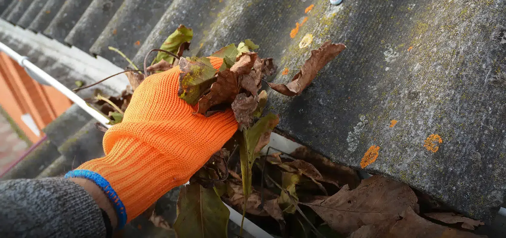 A person wearing orange gloves is cleaning a gutter on a roof.