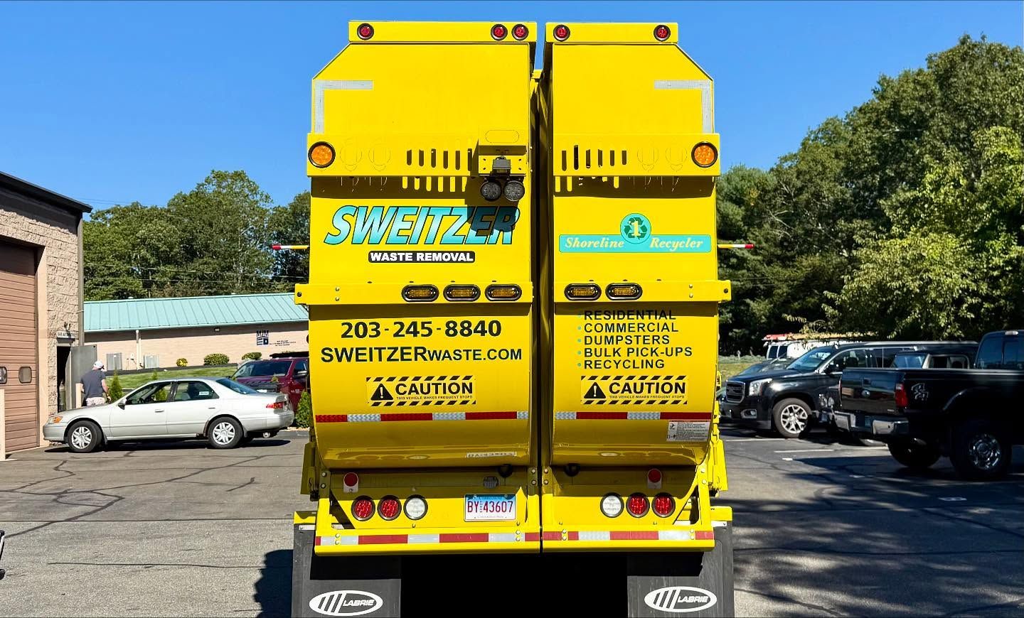 Rear view of a bright yellow garbage truck. Smeltzer Waste logo and contact info are visible. Blue sky in background.