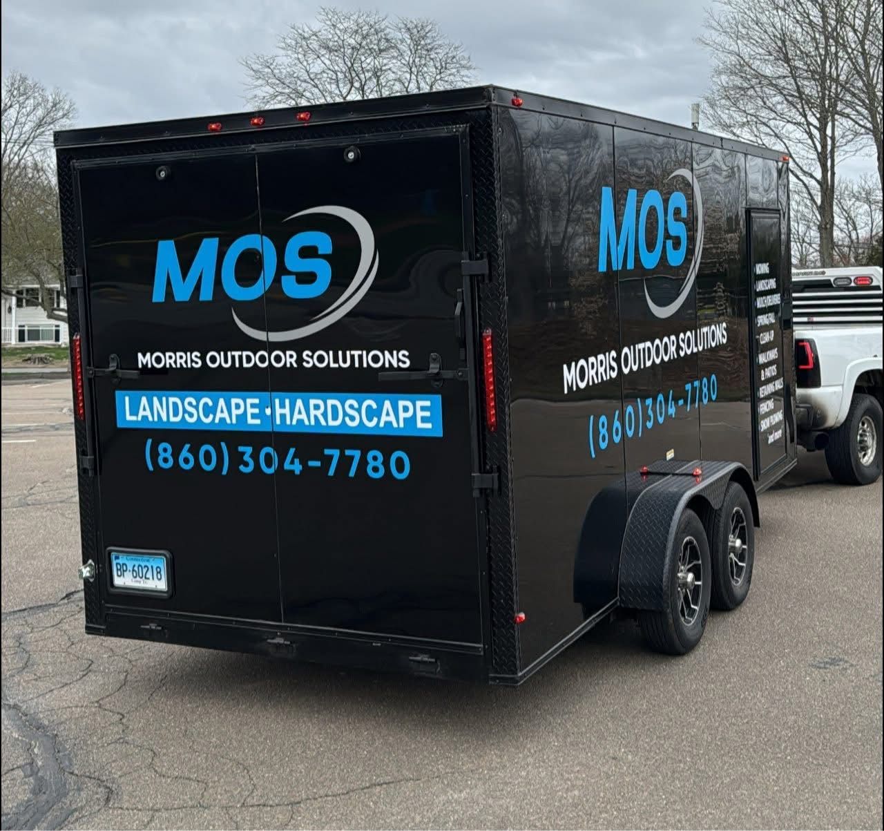 Black trailer with MOS logo and contact info, parked on pavement, hitched to a truck.