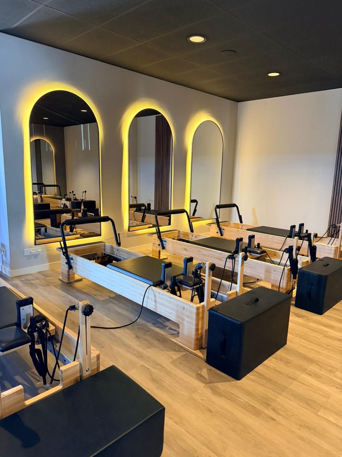 Pilatesstudio met reformer-apparaten, gebogen spiegels en warme gele accentverlichting.