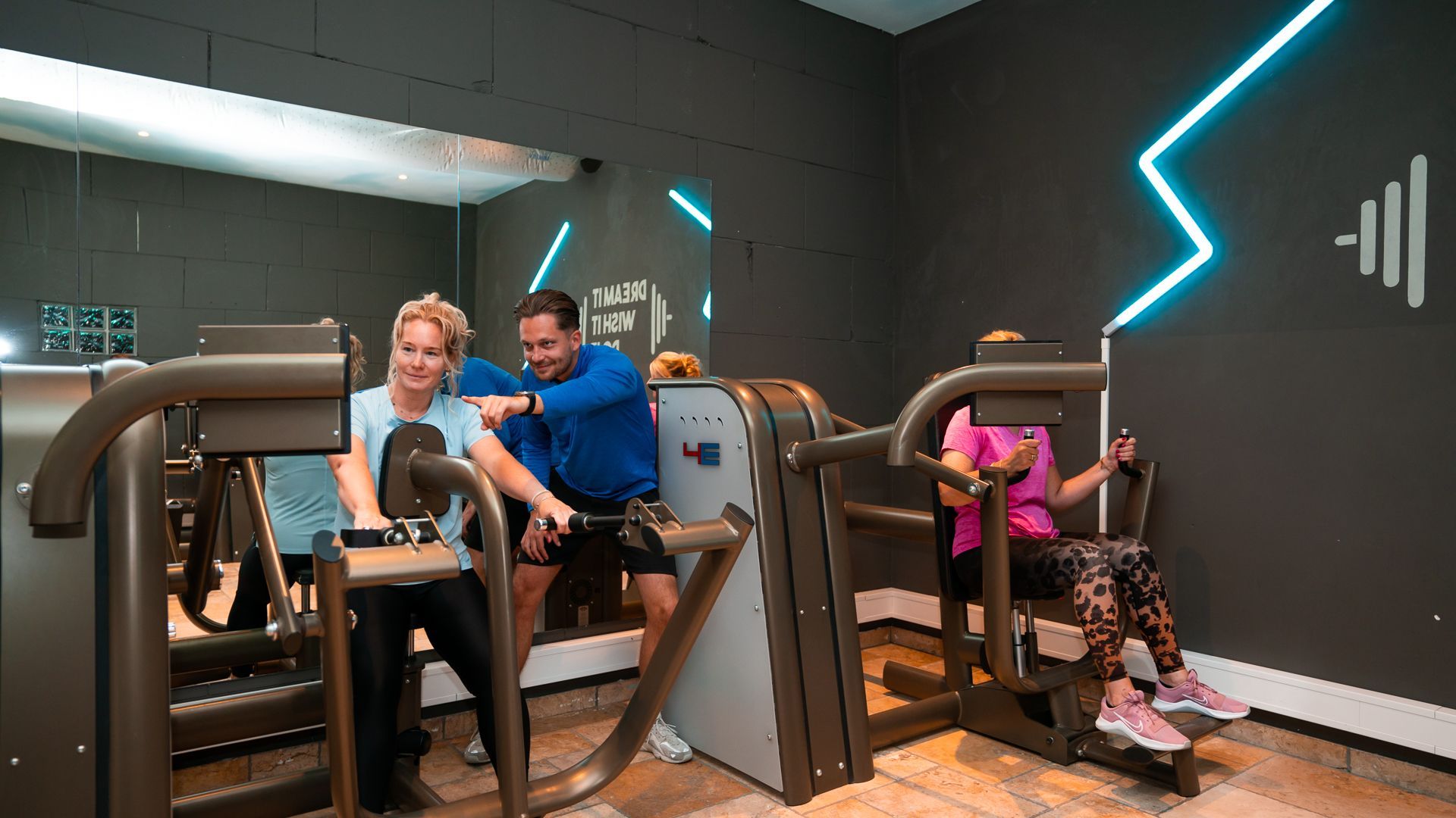 Twee mensen tillen halters in een sportschool, waarbij een van hen een gewicht boven zijn hoofd tilt naast een spiegel.