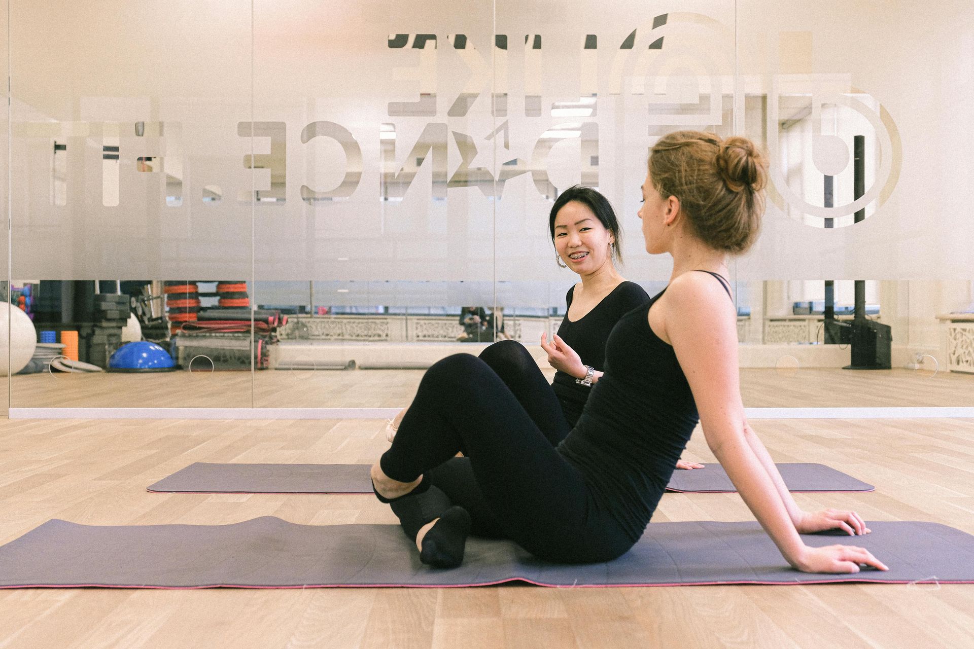 Twee mensen zitten op yogamatten in een studio, een glimlacht en de ander doet rek- en strekoefeningen naast haar.