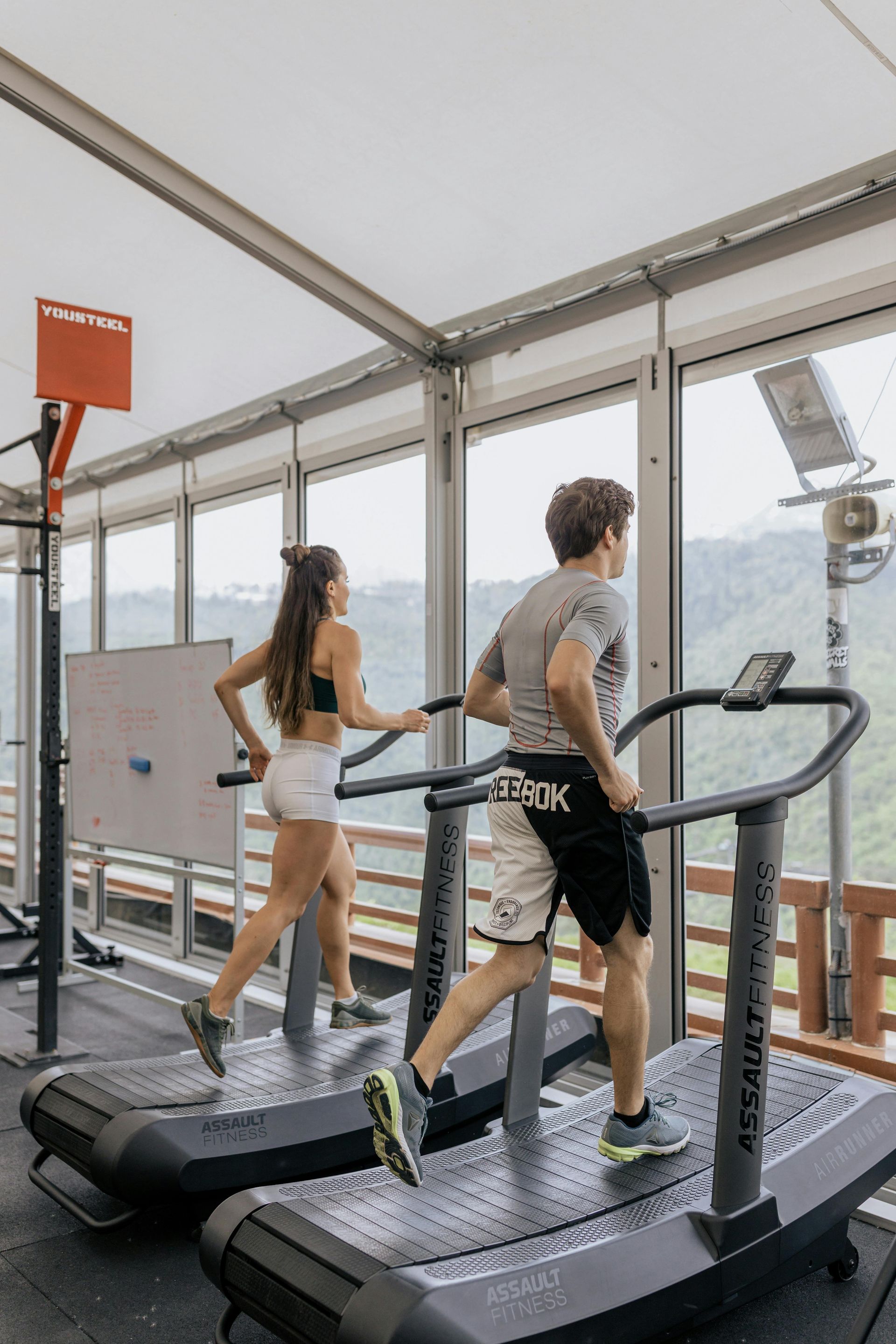 Twee mensen rennen op loopbanden in een sportschool met glazen wanden en uitzicht op de bergen.