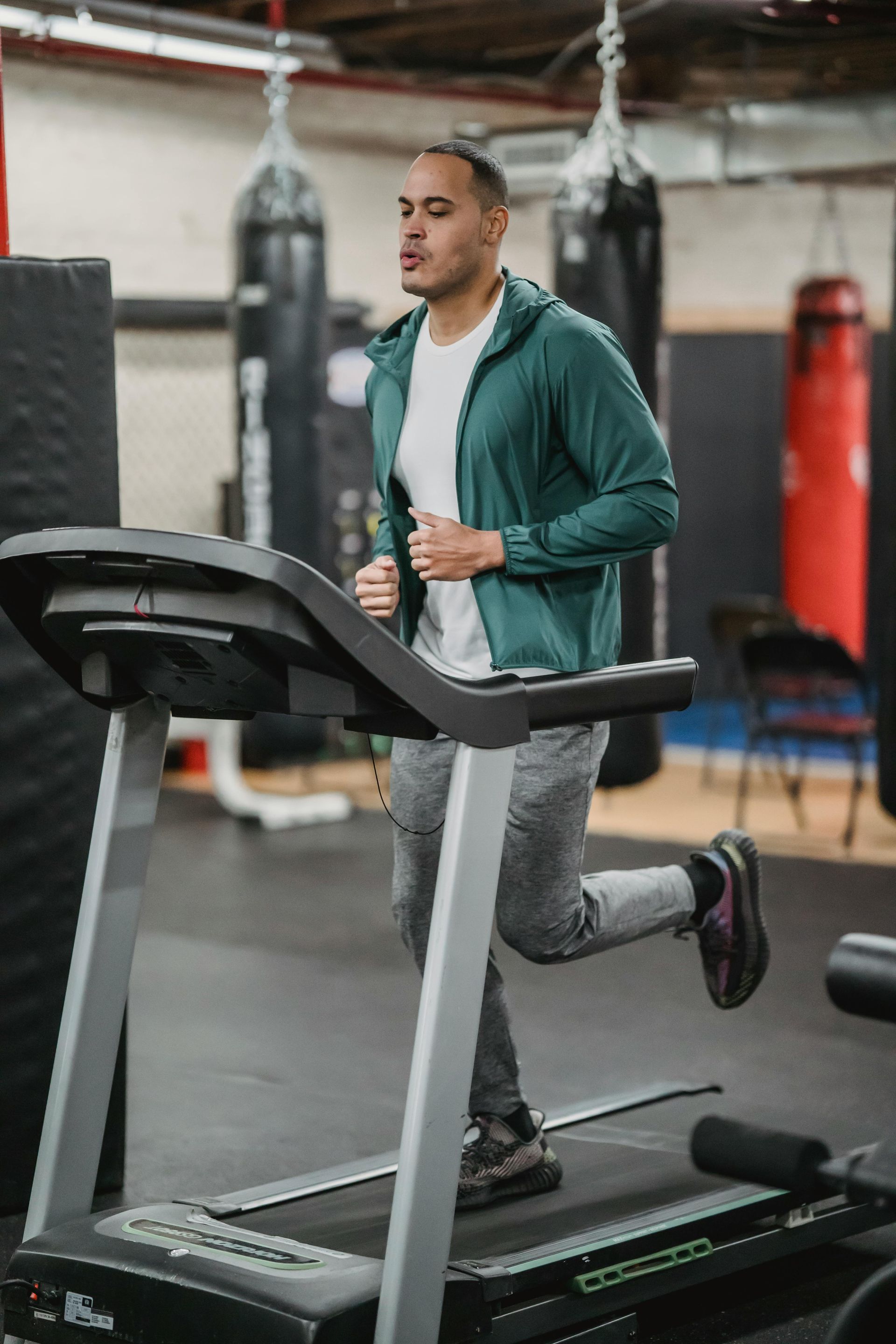 Een man jogt op een loopband in een sportschool, gekleed in een turquoise jas en een grijze broek.