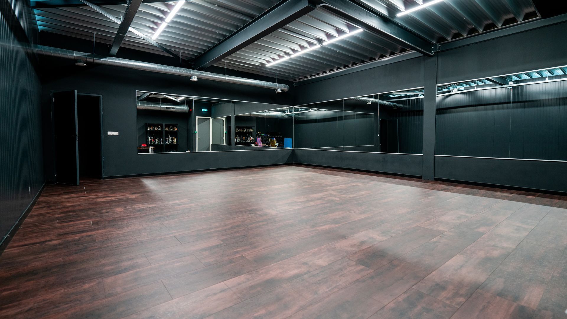 Een lege dansstudio met donkere muren, een spiegelende achterwand en een houten vloer onder fel plafondlicht.