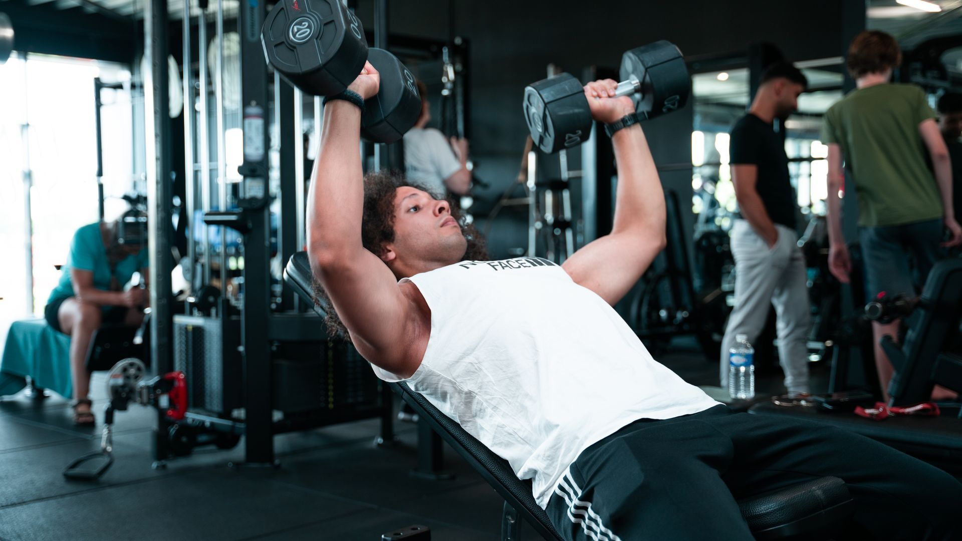 Een persoon doet een incline dumbbell press in een sportschool, met anderen en apparatuur op de achtergrond.