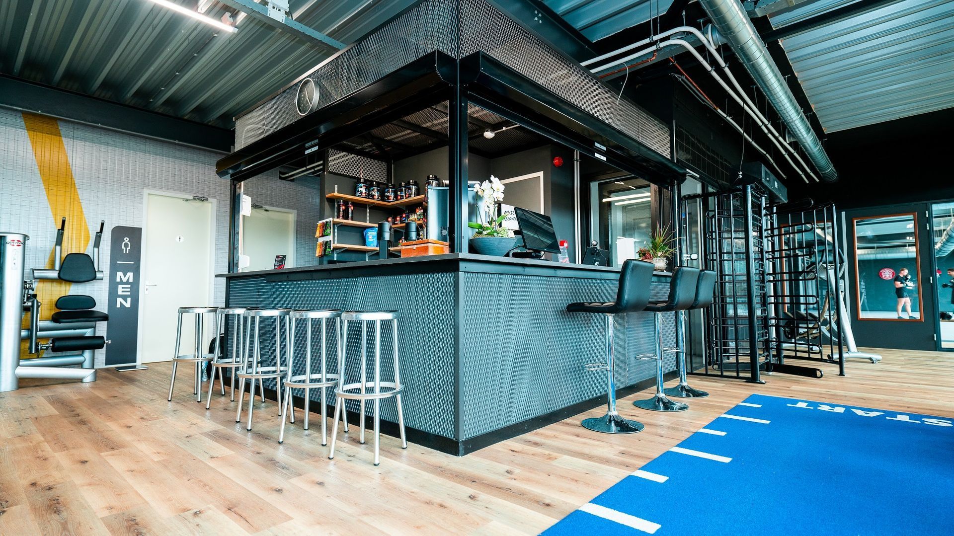 Modern barinterieur met metalen barkrukken, houten vloer en een blauw kunstgrasveld.