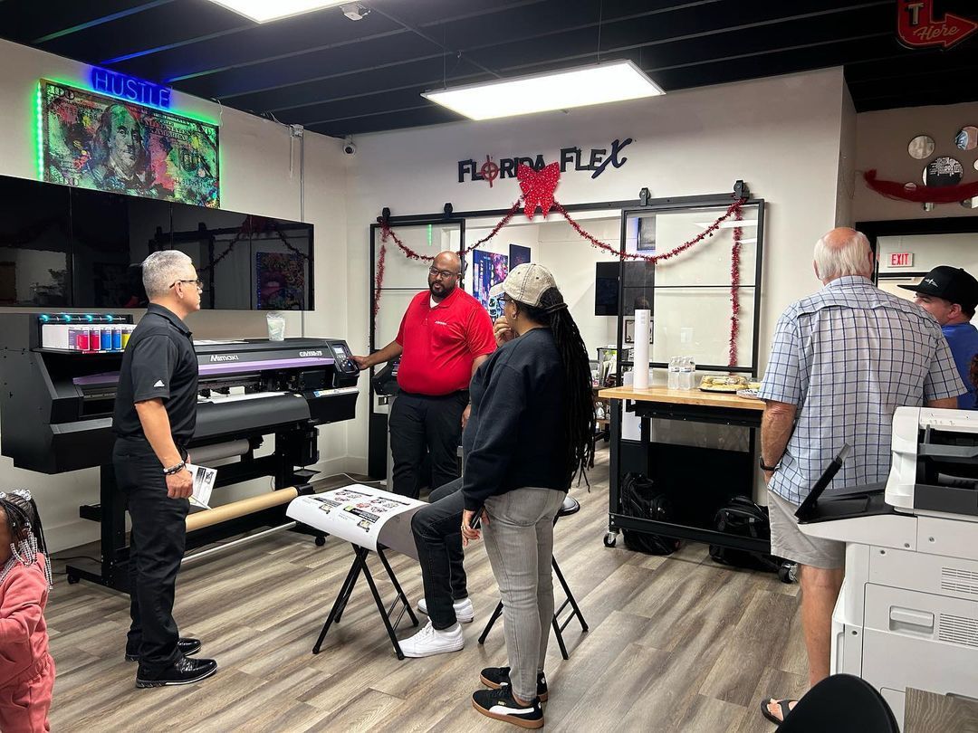 People in print shop with large printer, viewing printed material; man in red shirt explaining.