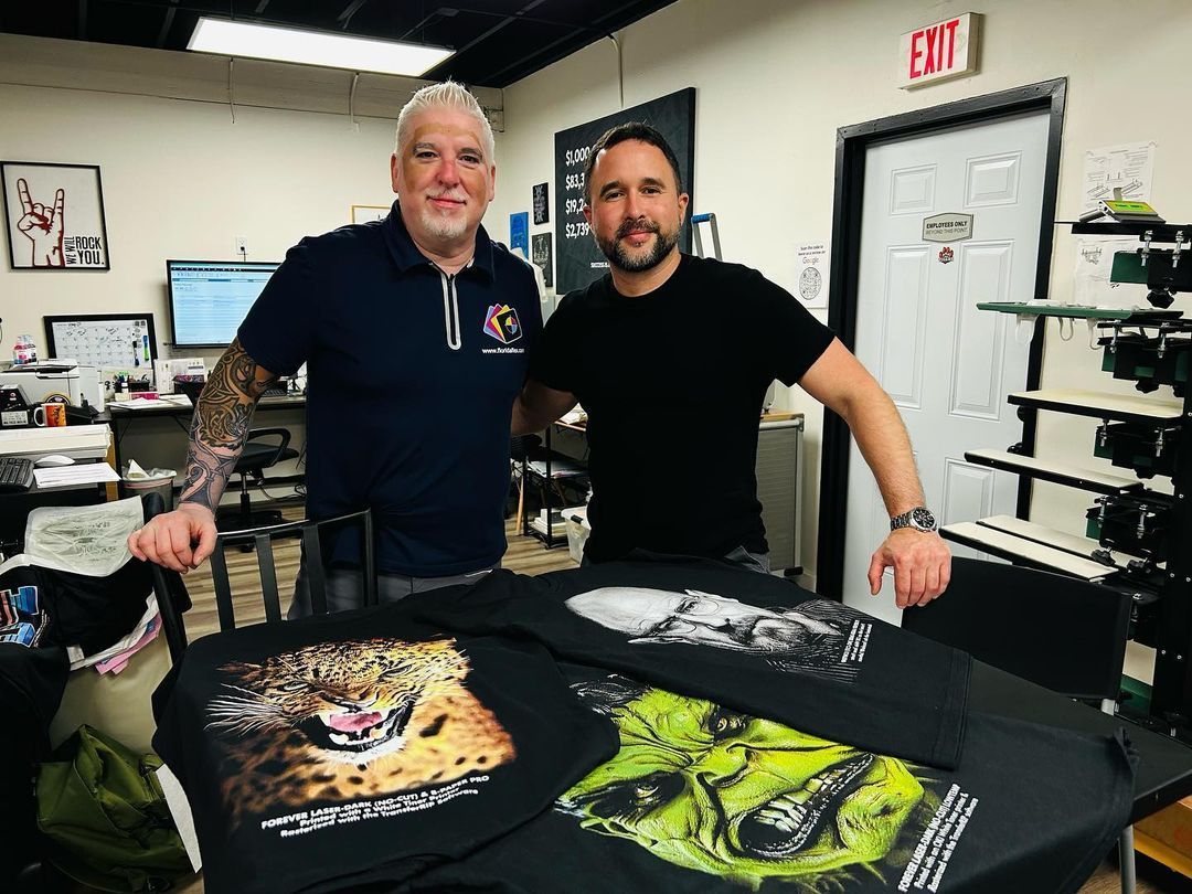 Two men stand near printed t-shirts in a print shop. One man has tattoos and the other has a beard.