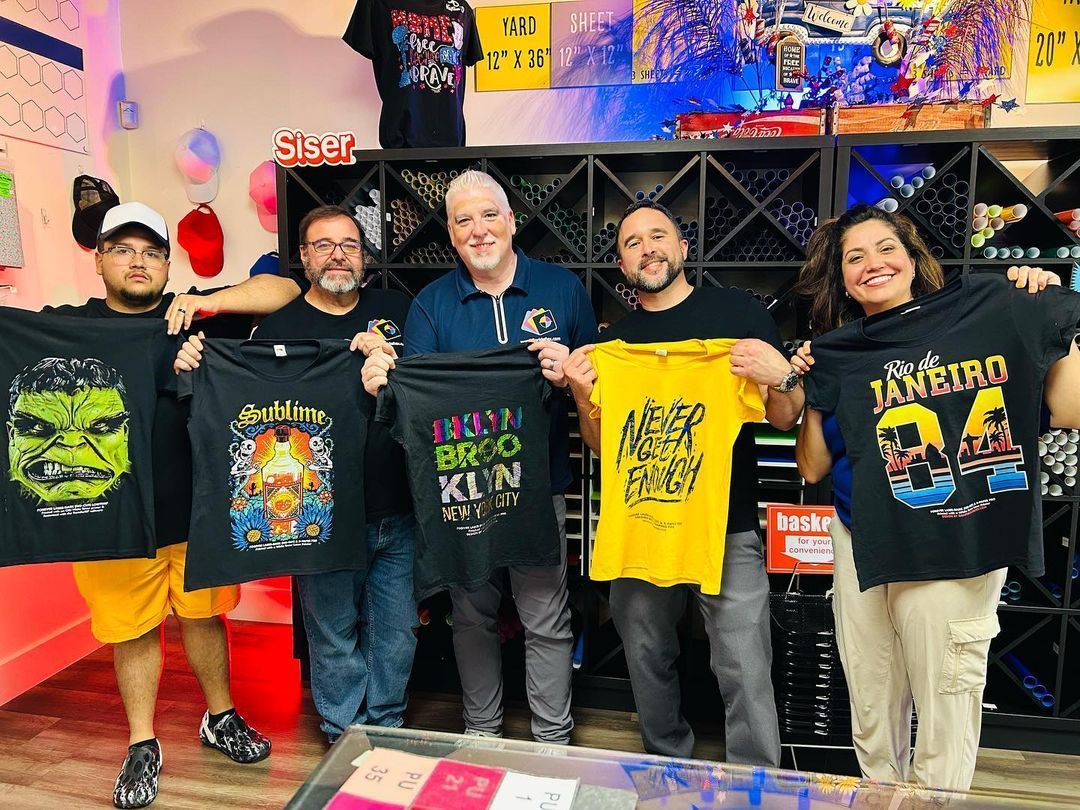 Six people holding up t-shirts with graphics, smiling in a shop with shelves.