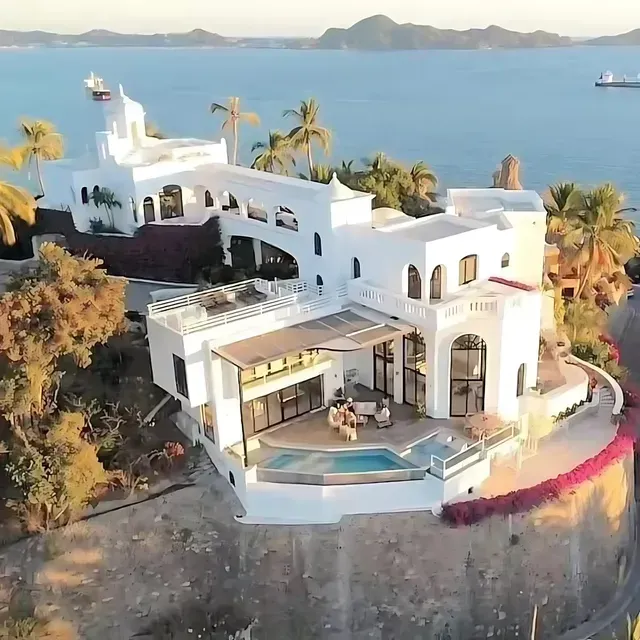 White villa perched on a cliff overlooking the ocean, palm trees, and small pool.