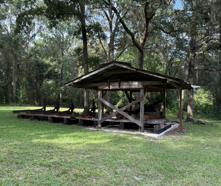 Wooden structure with a covered roof, in a grassy area with trees in the background.