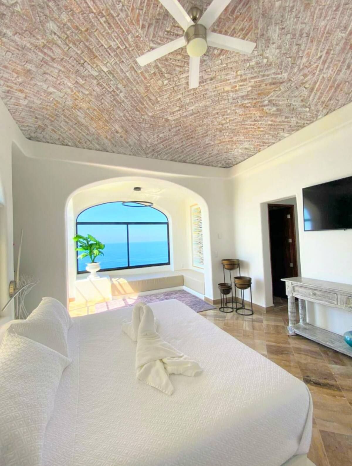 Bedroom with brick ceiling, large window overlooking the ocean, white bed, TV, and decorative table.