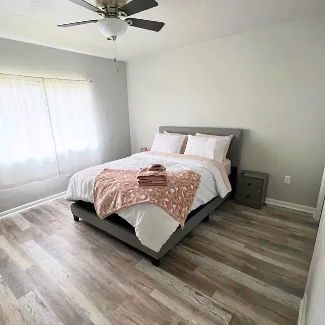Bedroom with a bed, nightstand, ceiling fan, and window. Gray walls, light wood flooring, and pink accents.