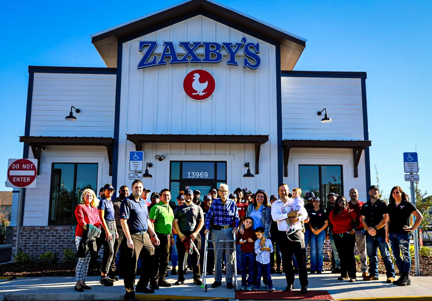 Zaxby's restaurant exterior with a crowd of people. Blue and white facade with a red chicken logo. People smiling.