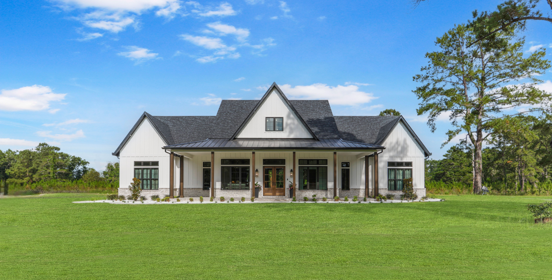 White farmhouse with black roof and porch, on a grassy lawn with trees and a blue sky.