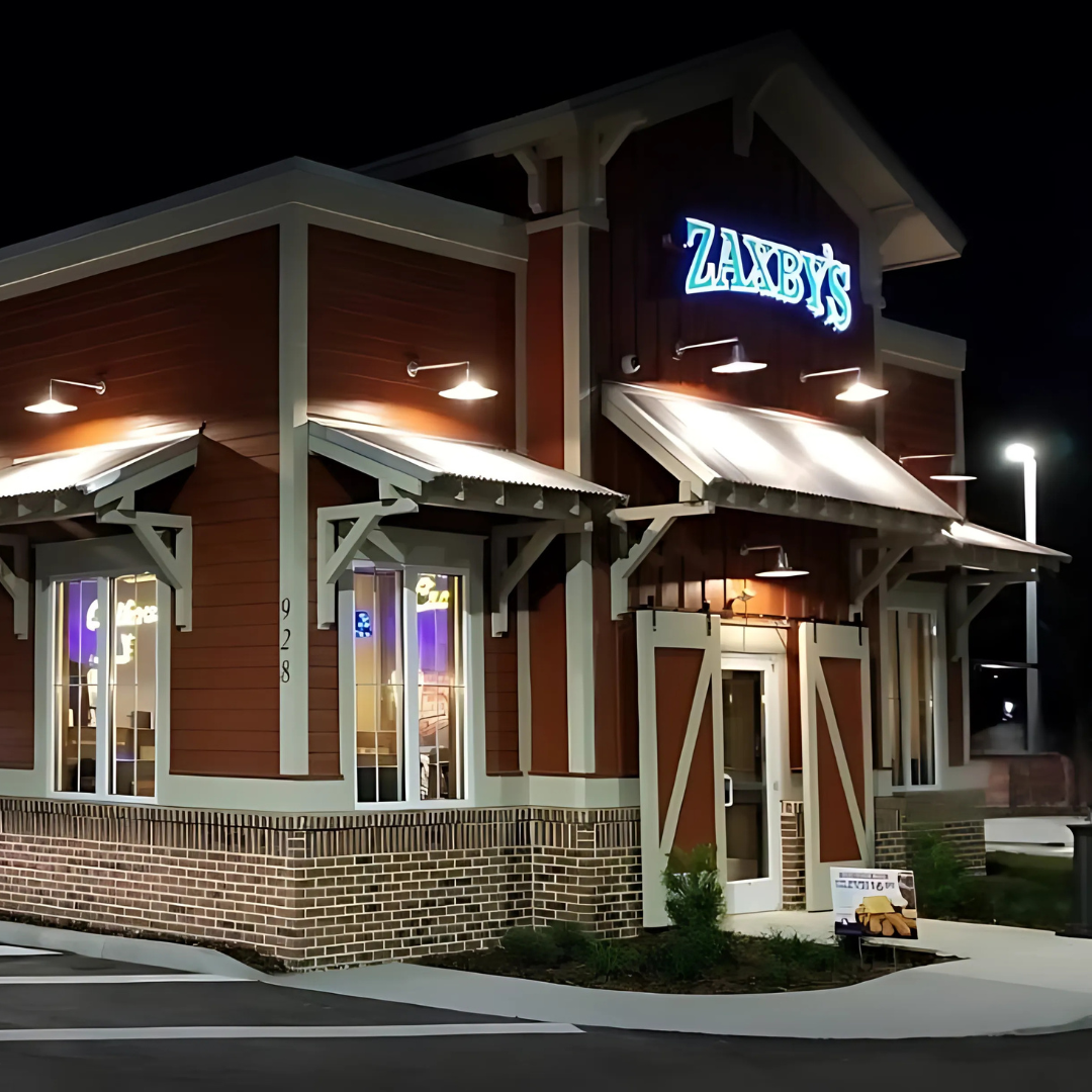 Zaxby's restaurant exterior at night with illuminated sign. Red siding, white trim, and brick base.