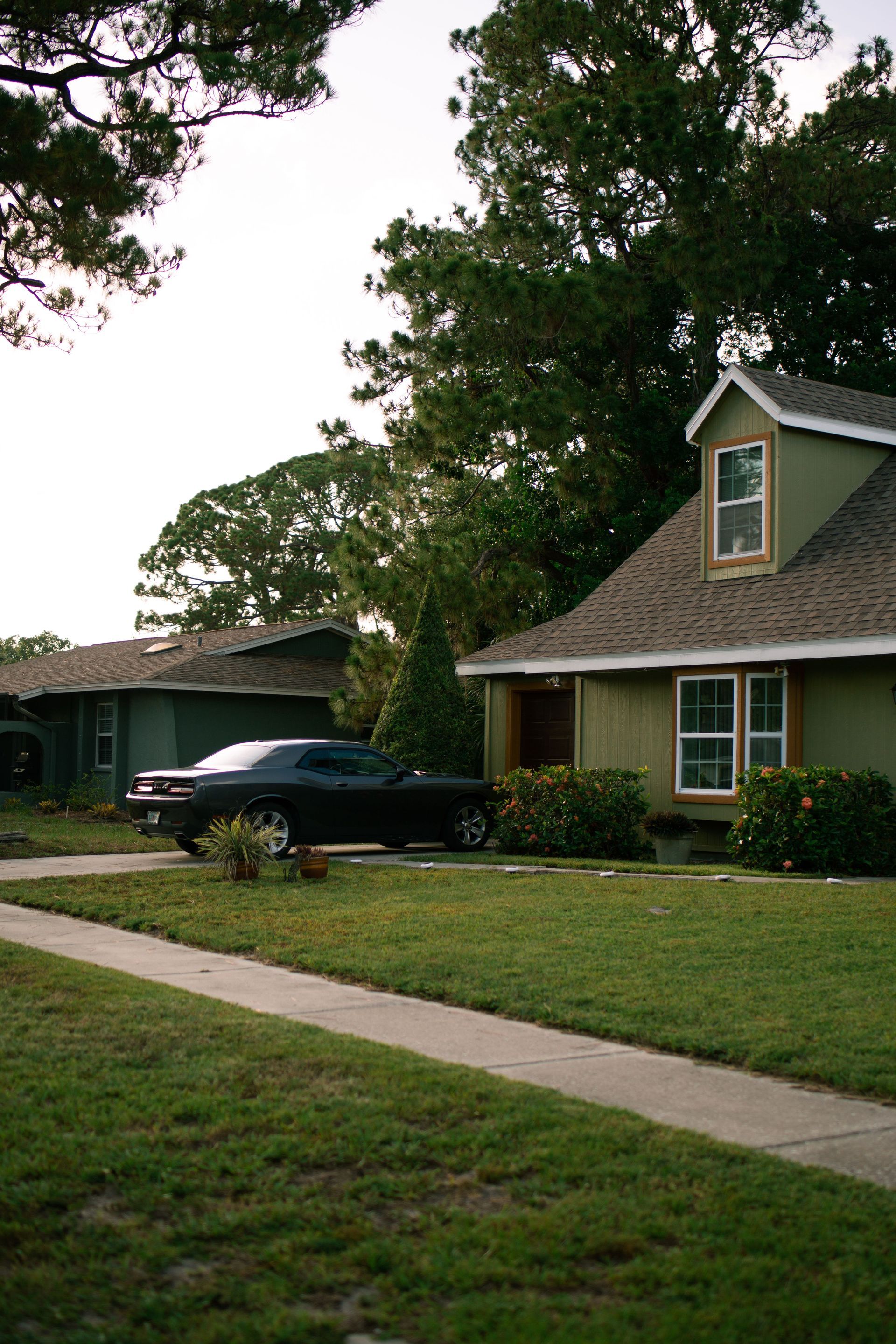 Suburban house with a dark car in the driveway, front lawn, and large trees overhead