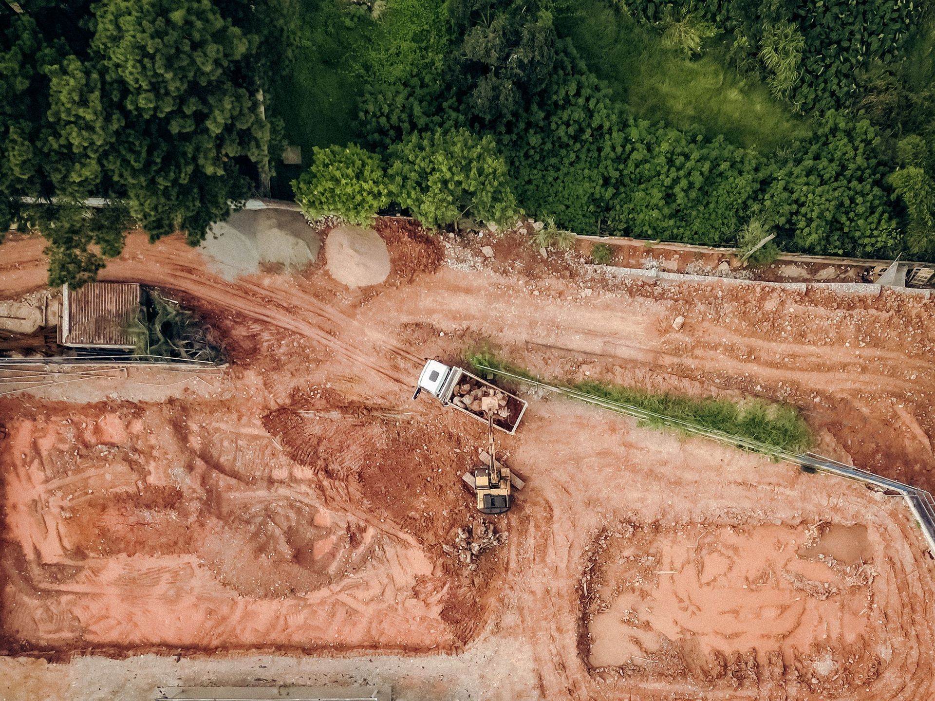 Aerial view of a muddy construction site with a dump truck and excavator on red soil near dense green trees