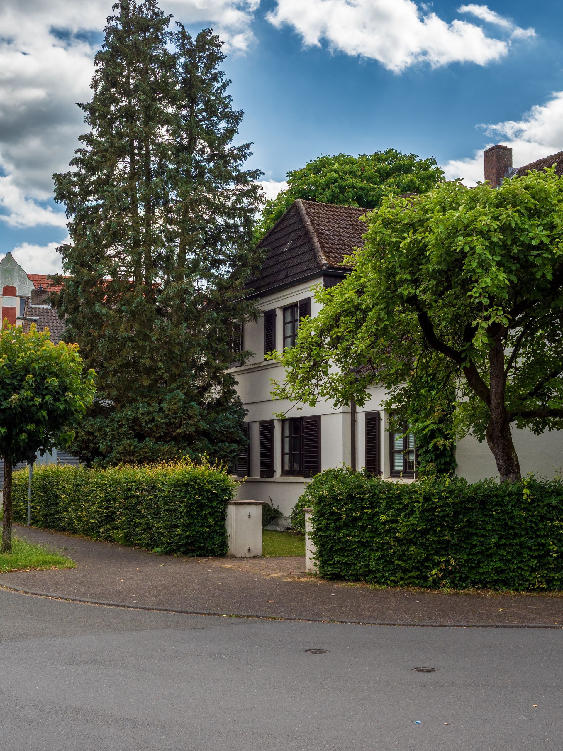 Quiet residential house with white walls, dark roof, and hedges beside a tree-lined street