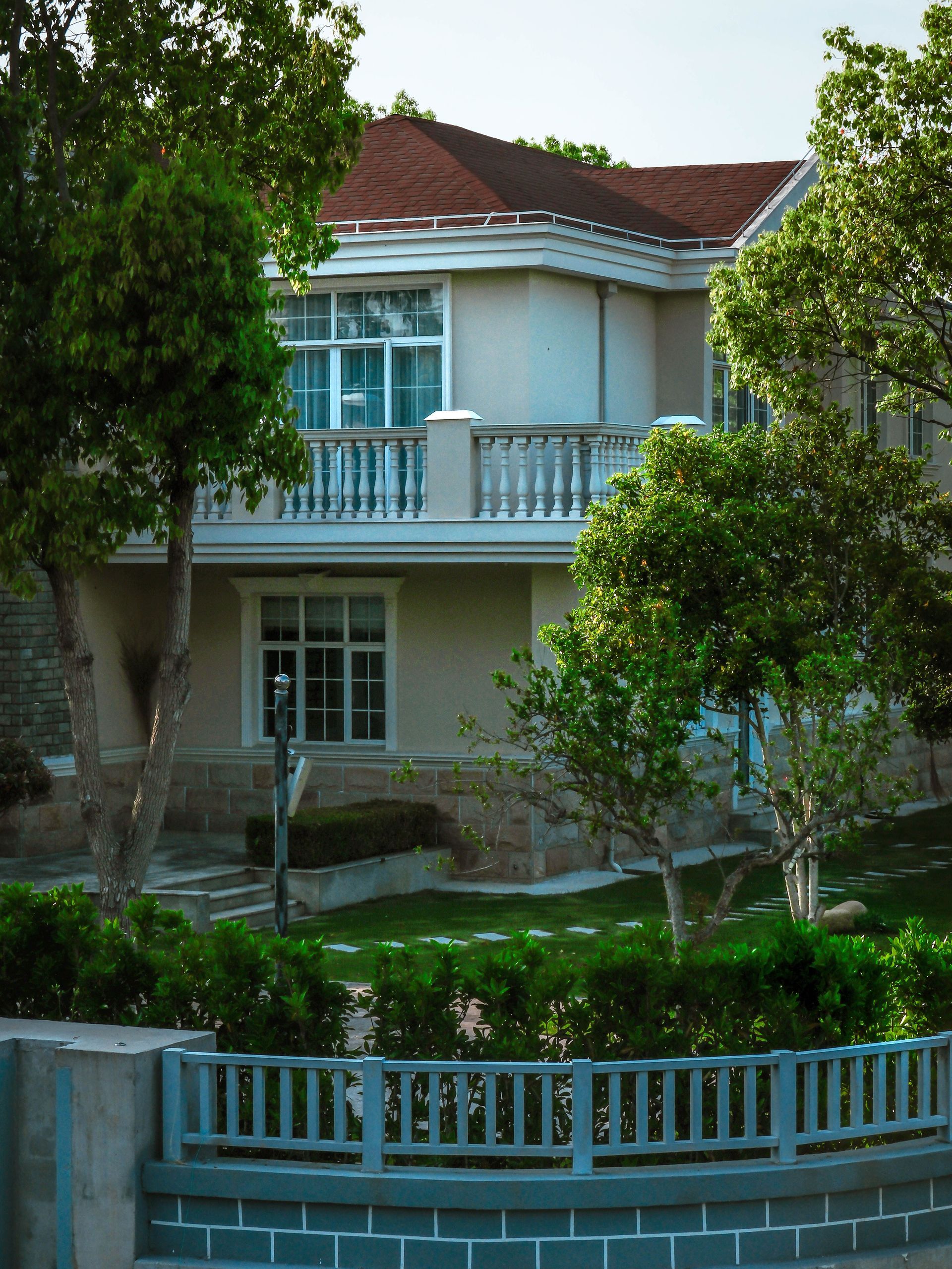 Two-story house with white walls, blue balcony railings, and red roof behind lush green trees.