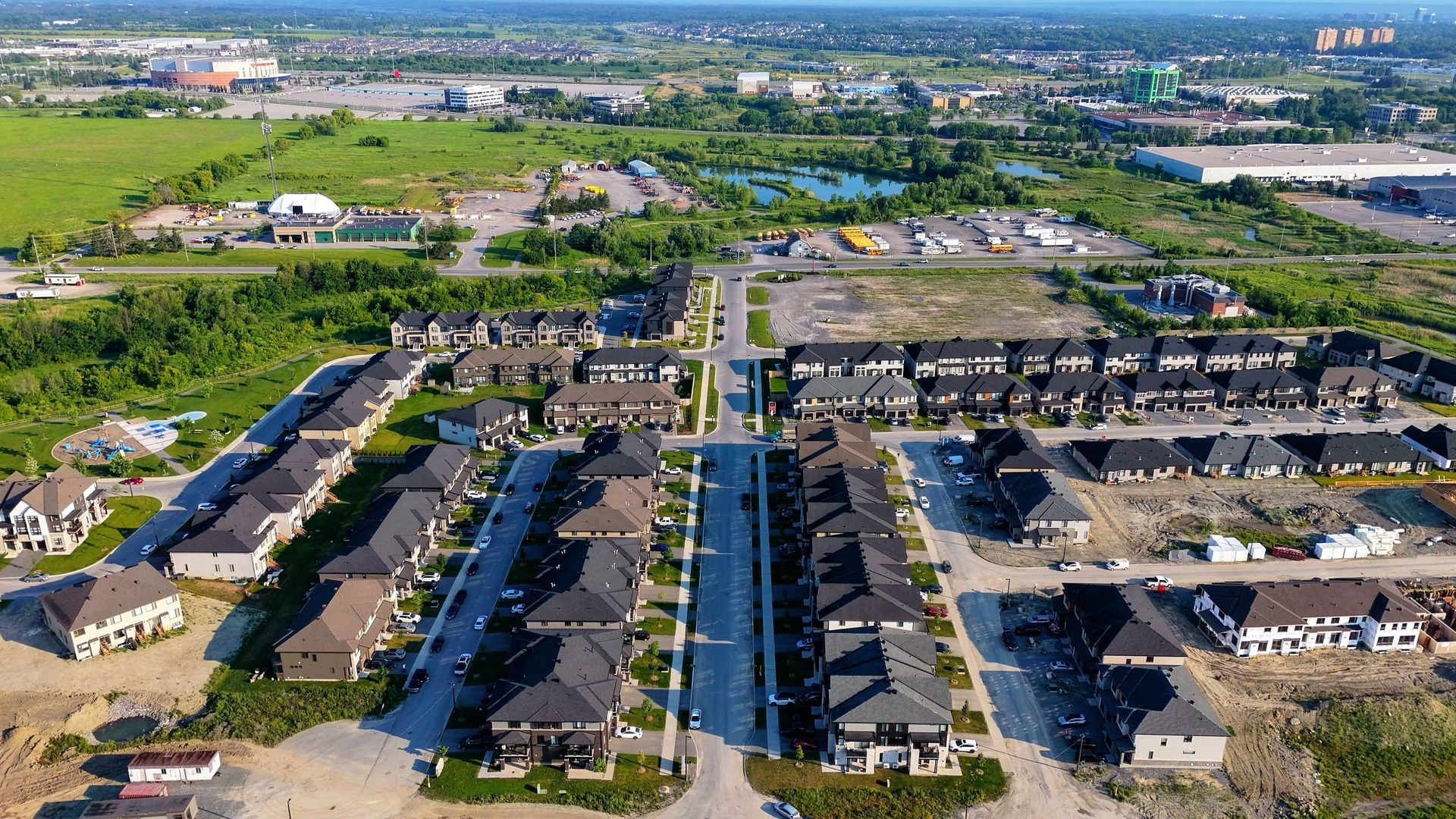 Aerial view of a suburban neighborhood with rows of houses, roads, and green fields in the distance.