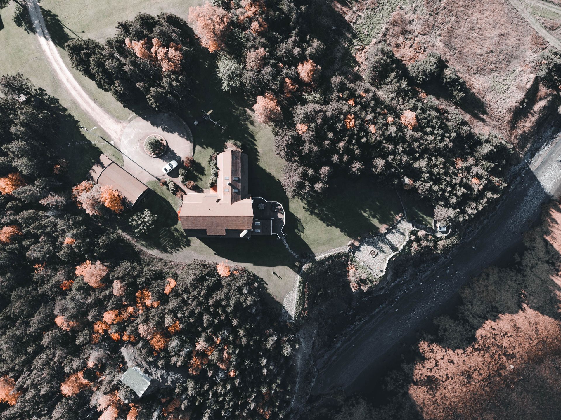 Aerial view of a house with a driveway, surrounded by trees beside a road and dark riverbank