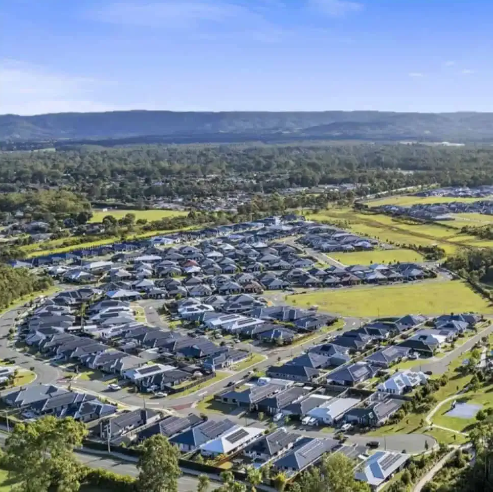 Aerial view of a suburban neighborhood with many houses, green spaces, and a forest backdrop.