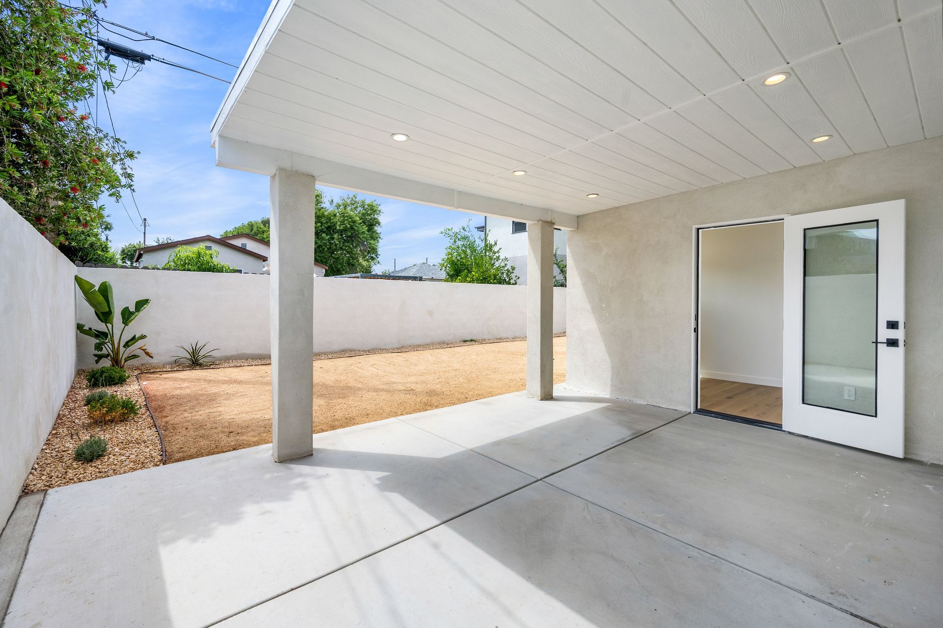 Covered concrete patio with white walls, open door, and gravel yard in bright daylight