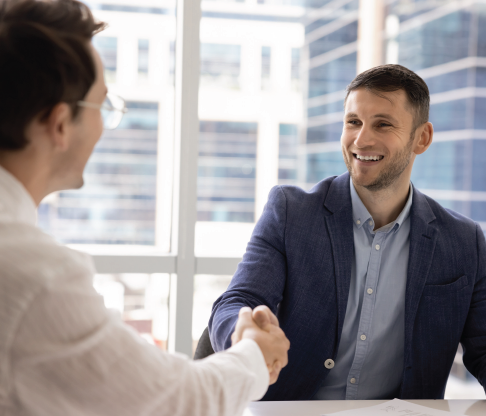 Two people shake hands across a table in a bright office with large windows.