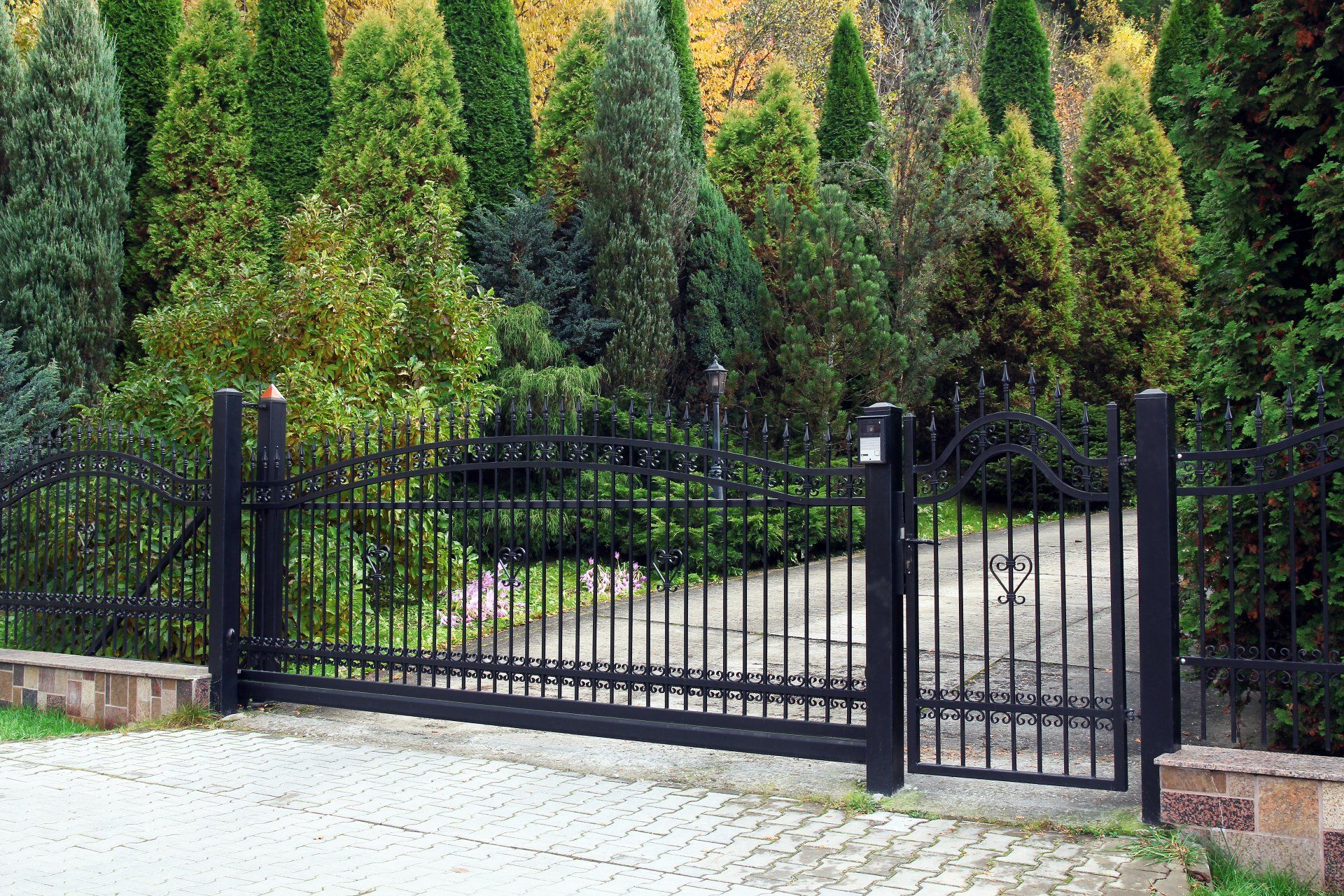A black fence is surrounding a driveway with trees in the background.