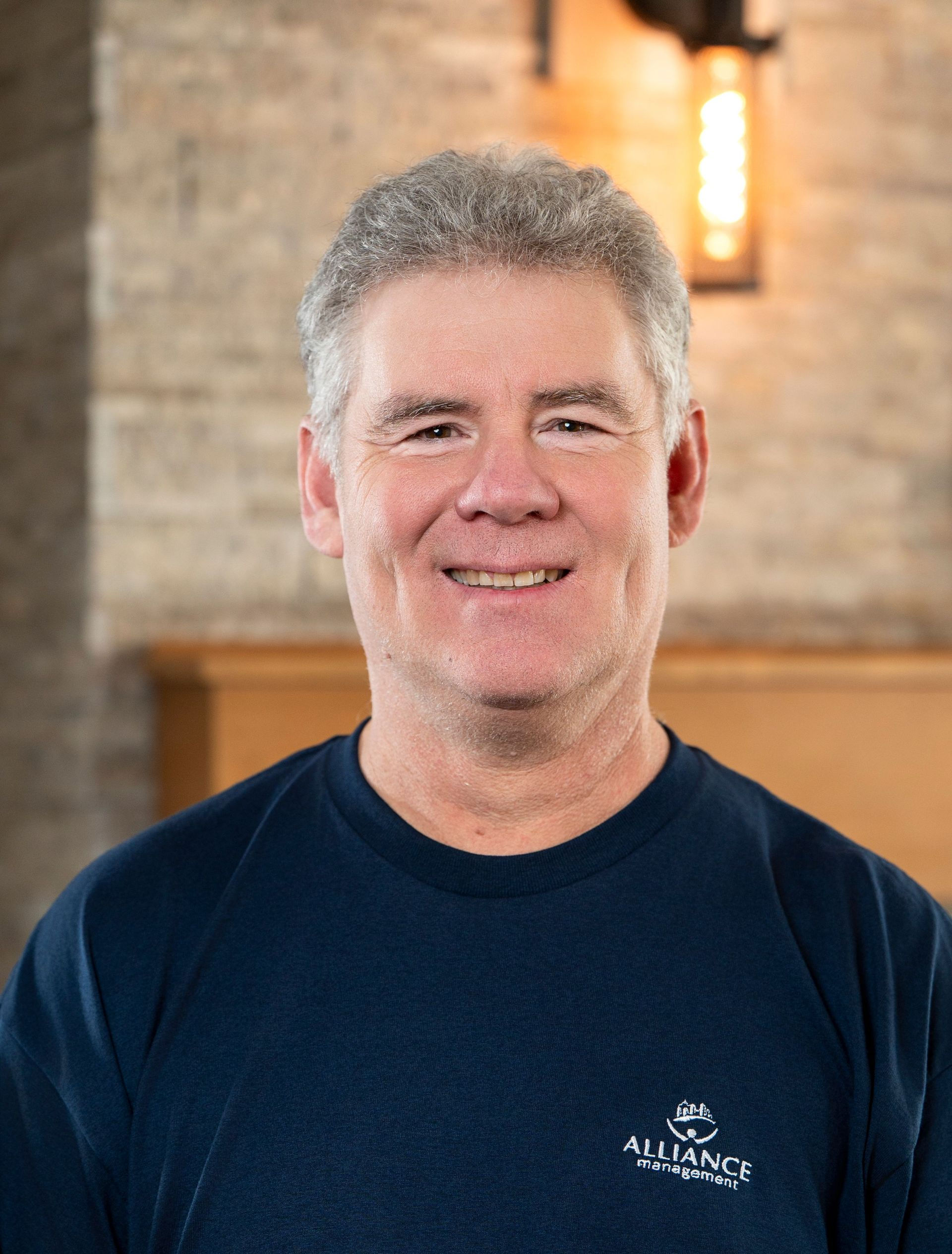 Portrait of a smiling man in a navy shirt, standing indoors against a warm brick background