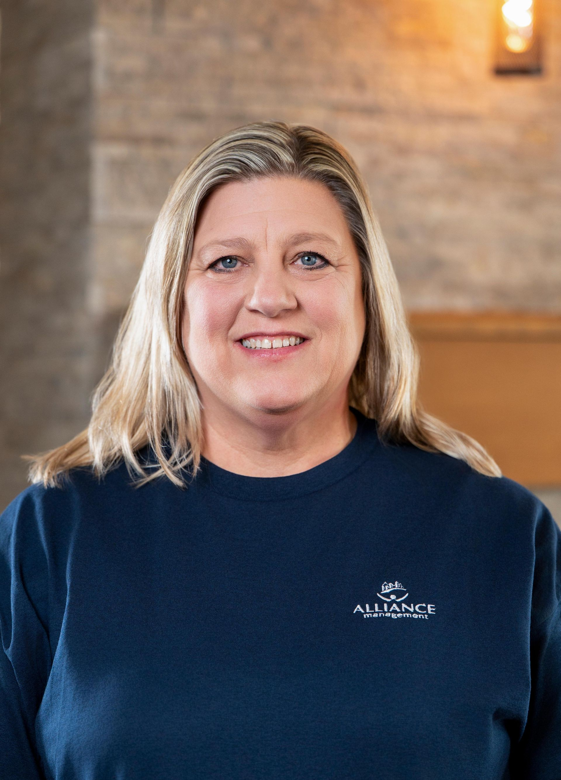 Portrait of a smiling woman in a navy sweater standing indoors against a blurred brick wall.