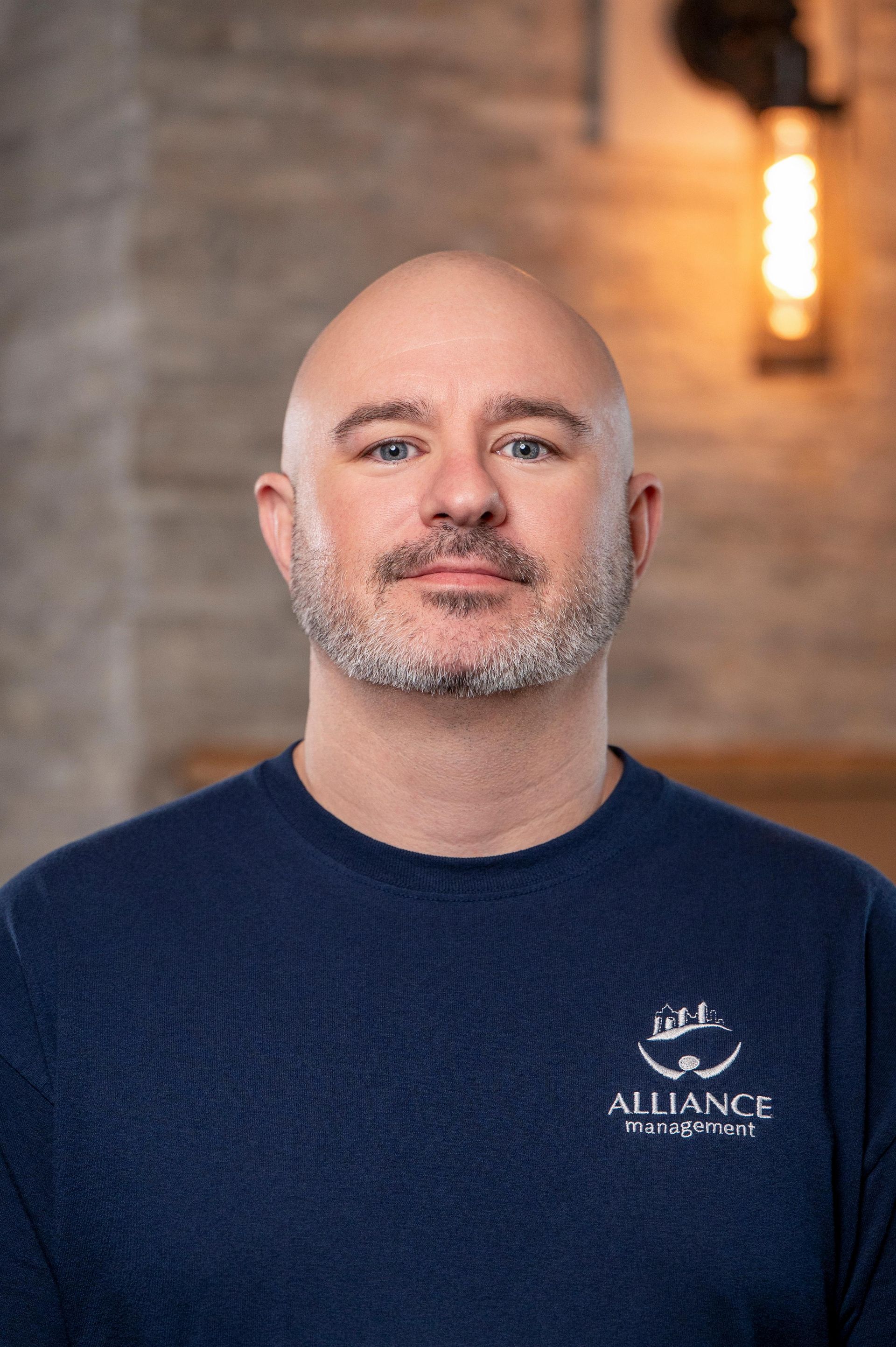 Portrait of a bald man in a navy shirt standing indoors against a brick wall with a warm light
