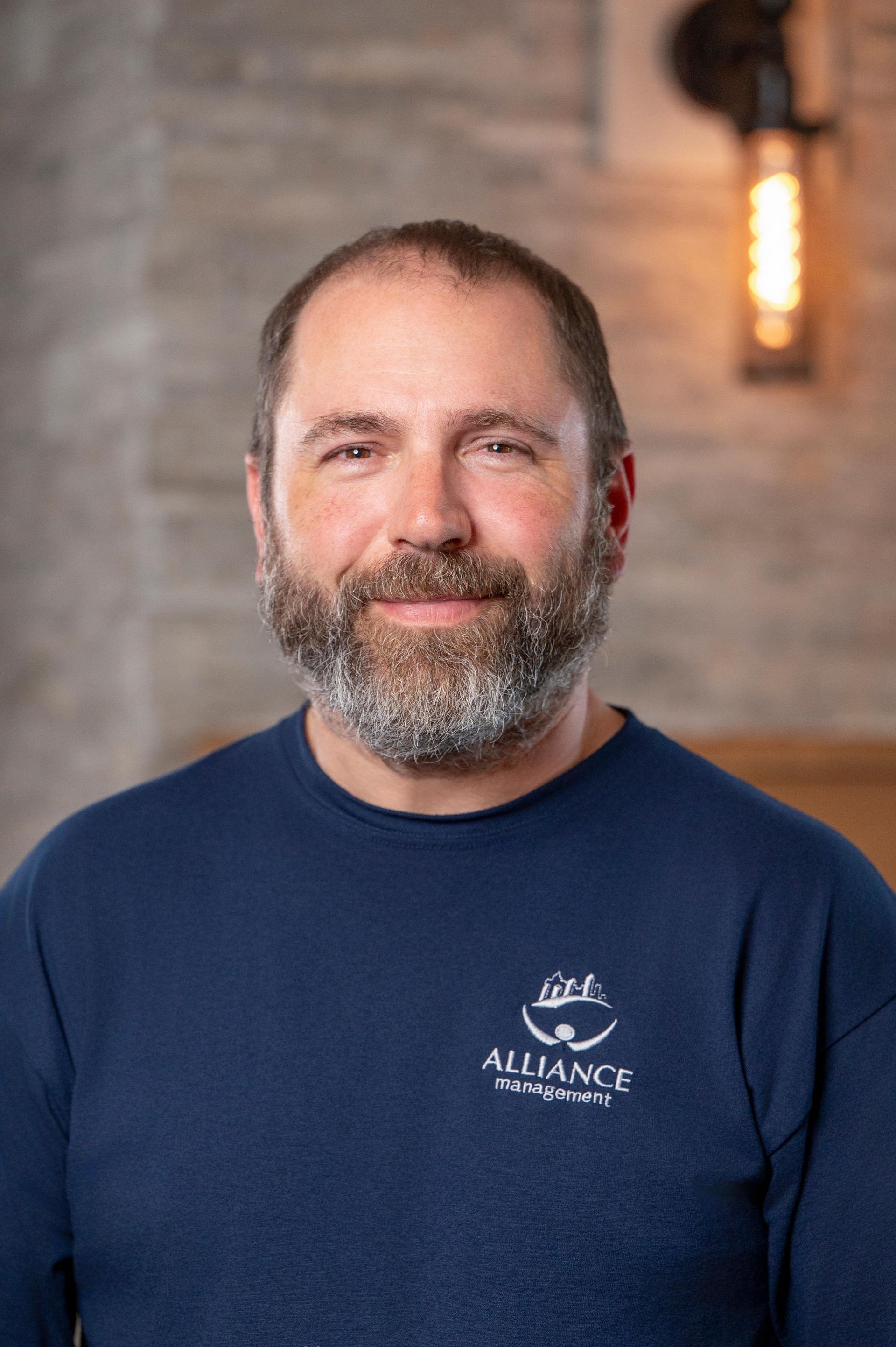 Smiling man with a beard in a navy shirt standing indoors against a brick wall.