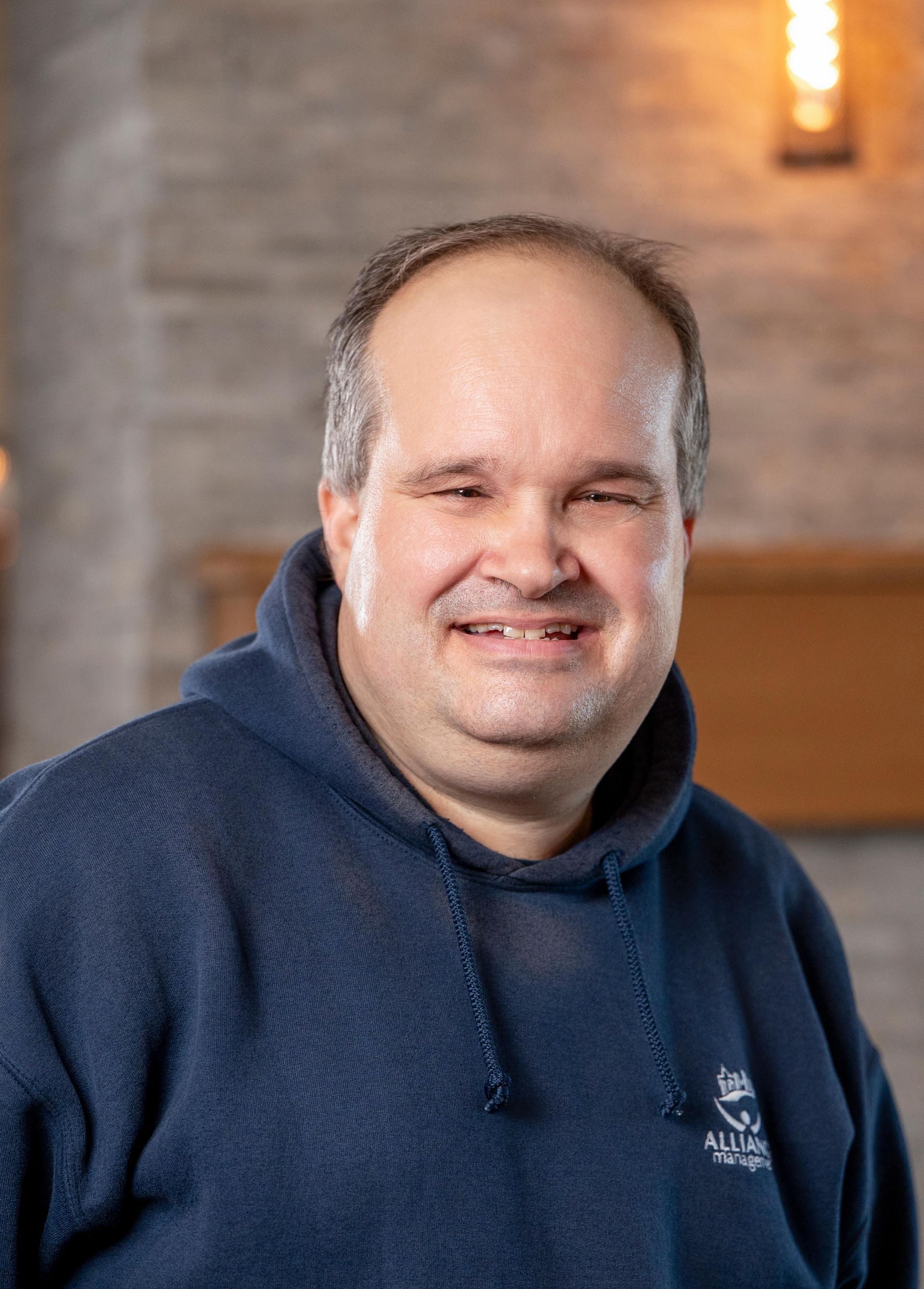 Smiling man in a navy hoodie standing indoors against a blurred brick wall background