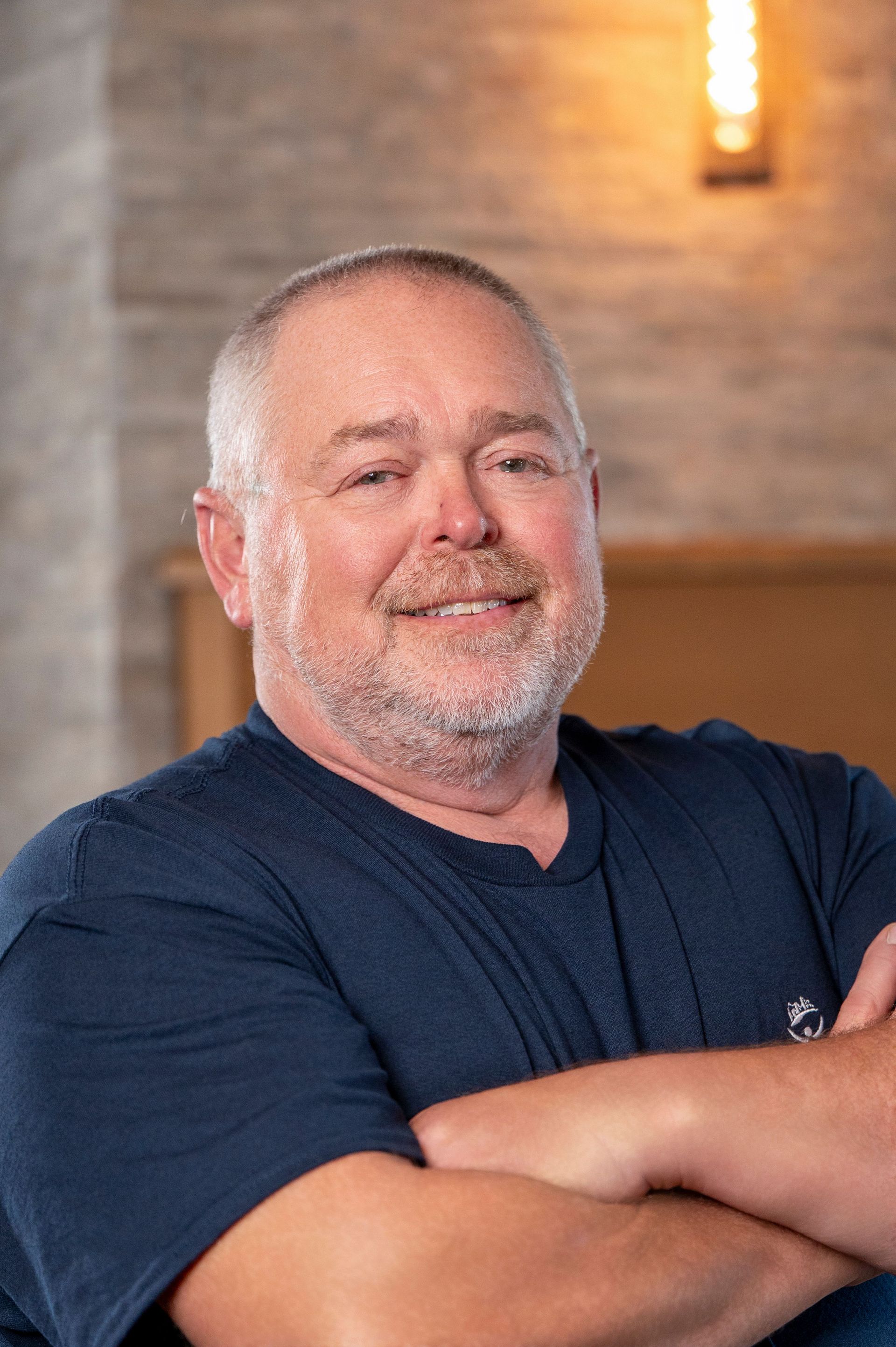 Man in a navy shirt smiling with arms crossed against a warm indoor brick wall background