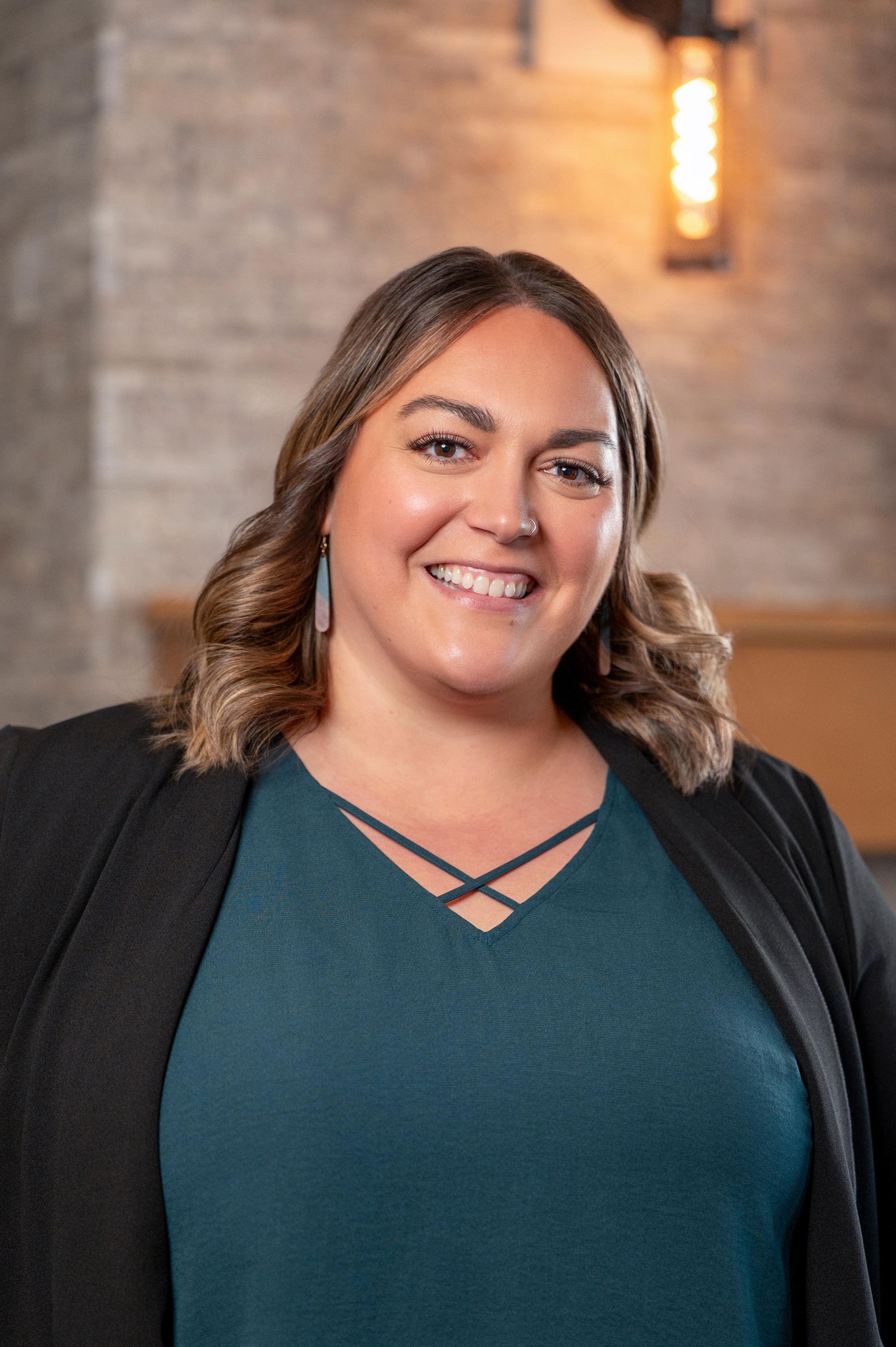 Smiling woman in a teal top and black cardigan standing indoors against a brick wall.