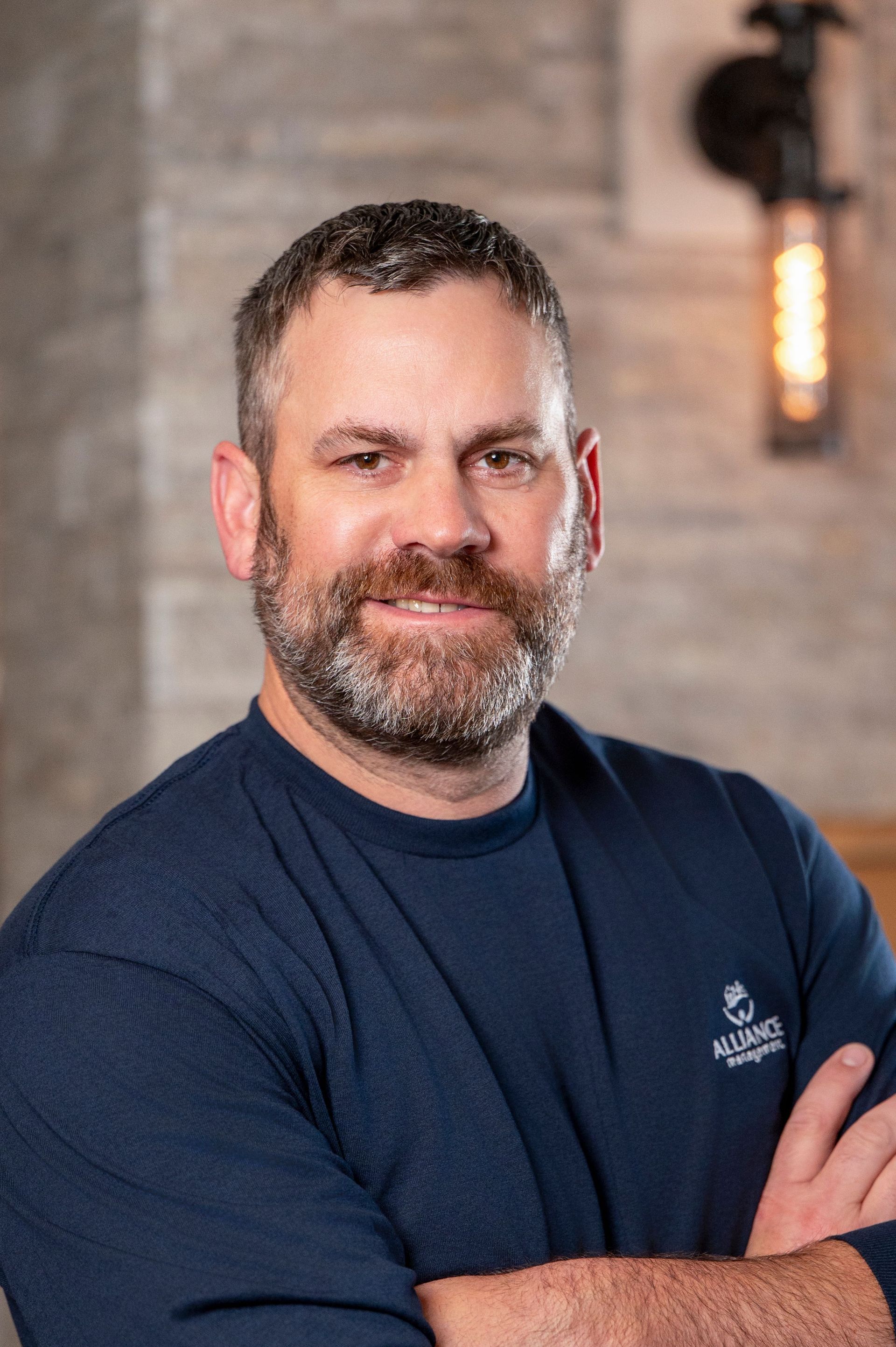 Portrait of a smiling man with crossed arms, wearing a navy shirt against a blurred indoor background.