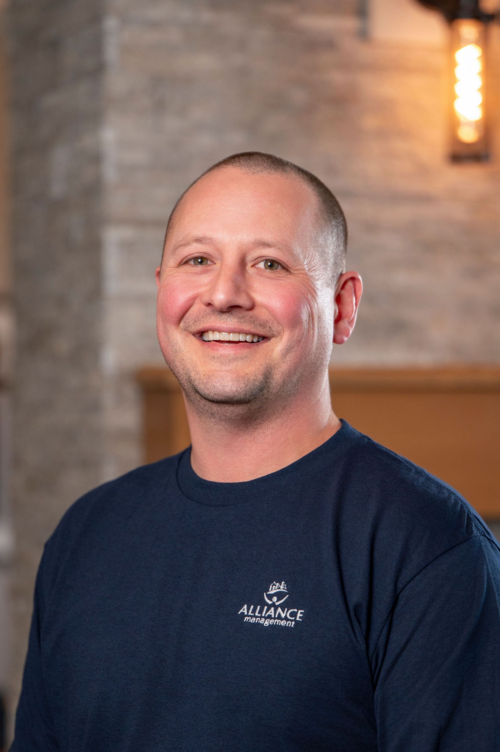 Smiling man in a navy T-shirt standing indoors against a brick wall background