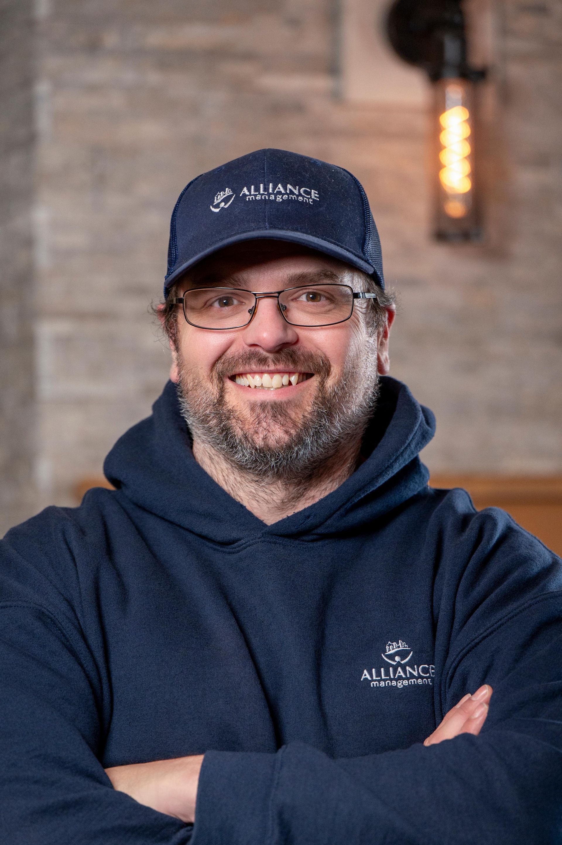 Smiling person in navy hoodie and cap, standing with arms crossed indoors.