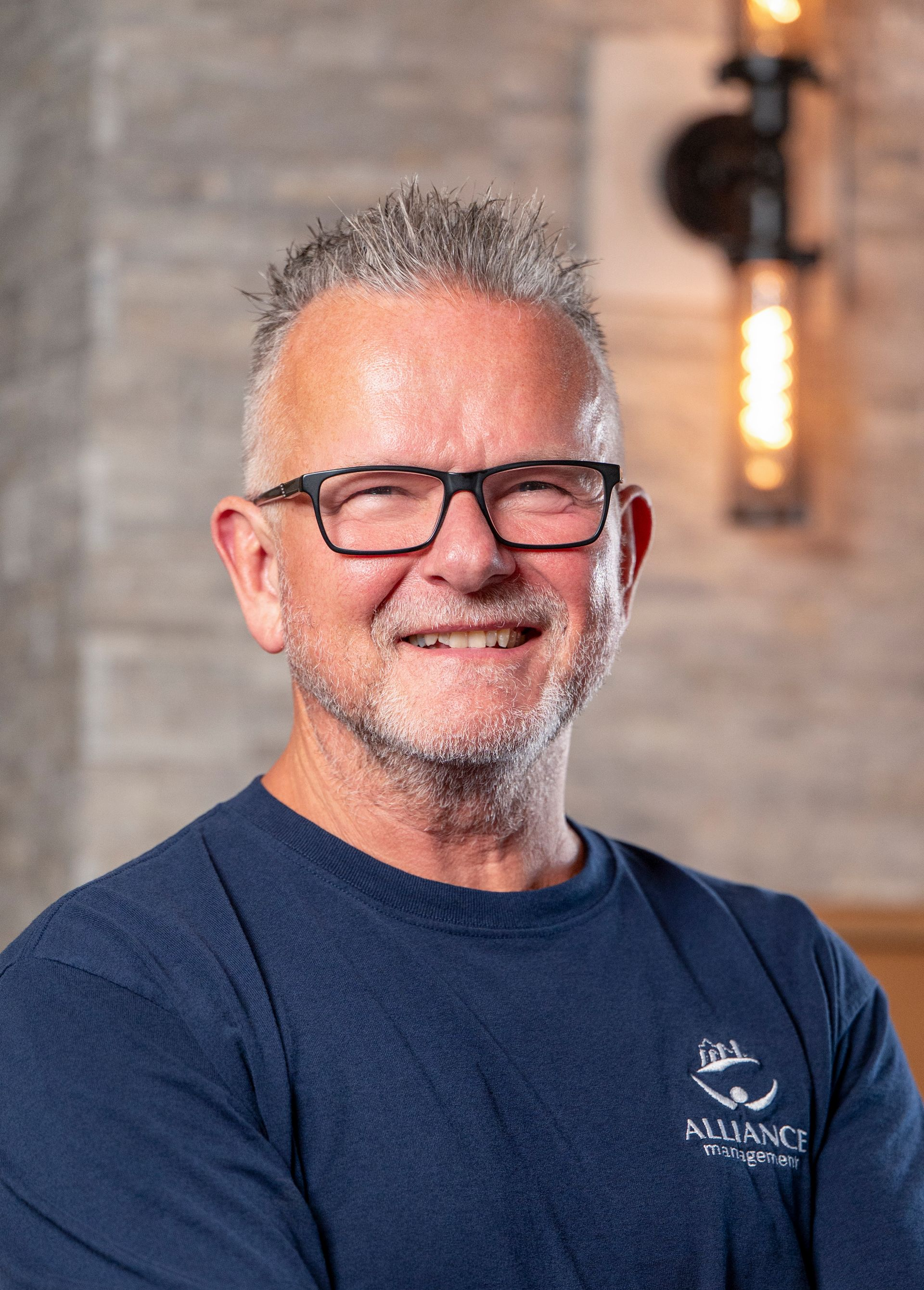 Smiling man in glasses wearing a navy shirt, standing indoors against a blurred brick wall.