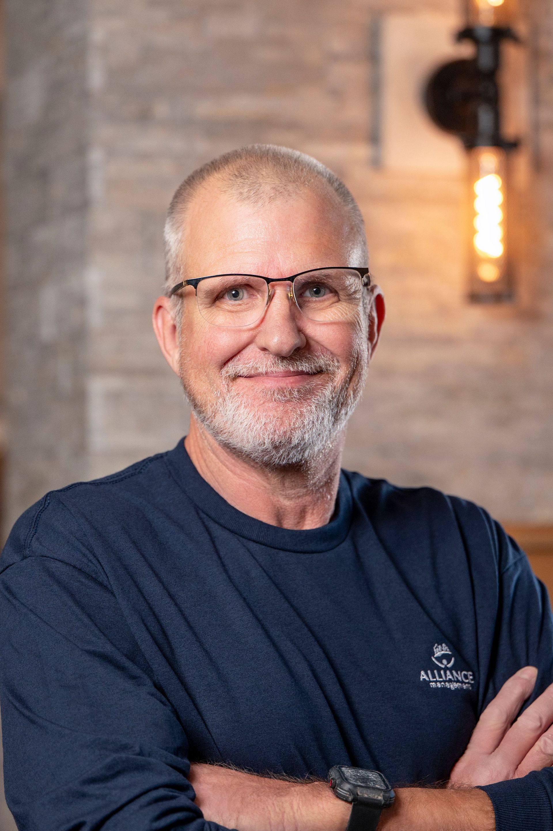 Smiling man with glasses and folded arms in a navy shirt against a warm indoor background