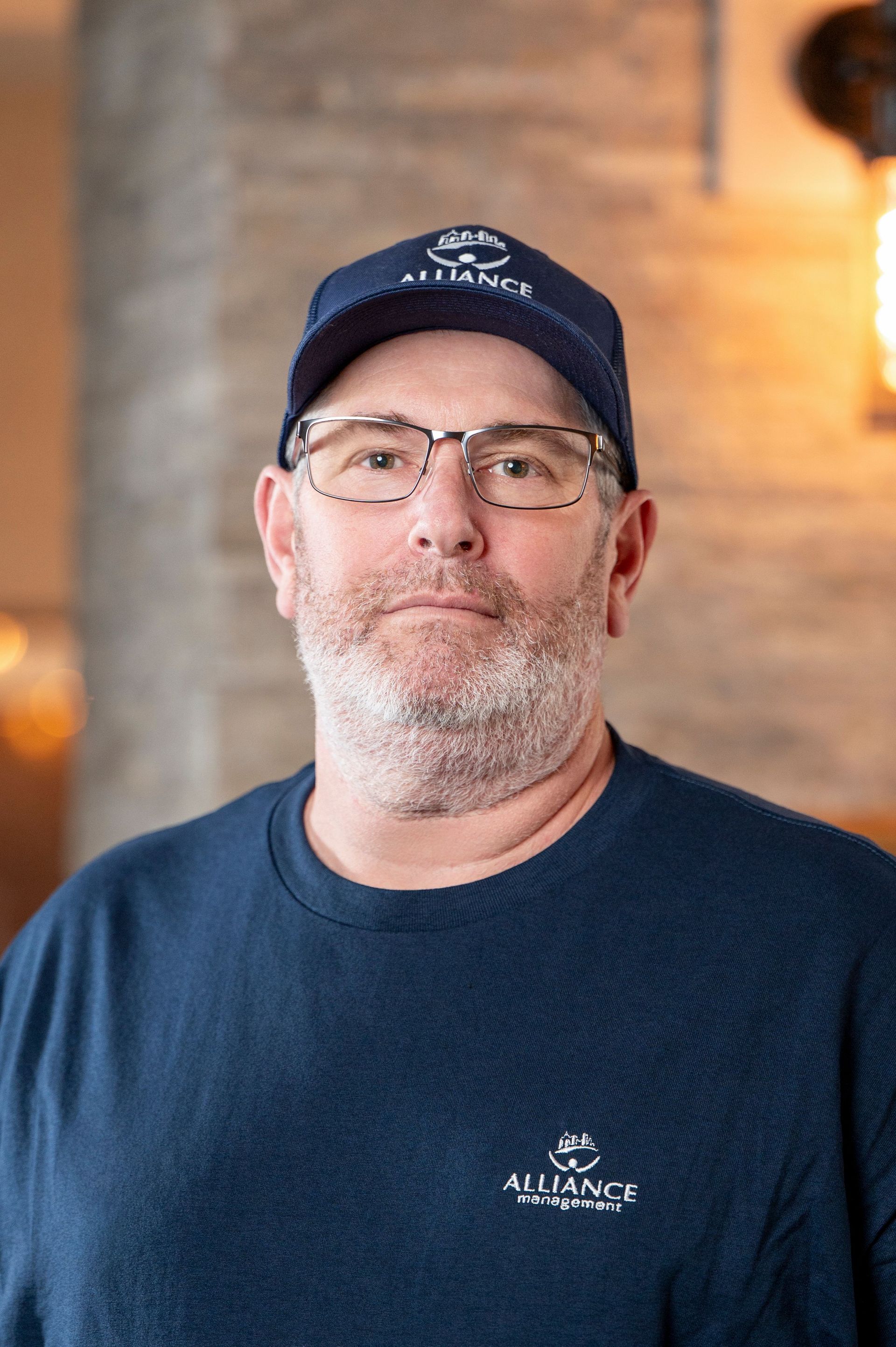 Man in navy cap and shirt, wearing glasses, standing indoors against a softly lit brick wall.