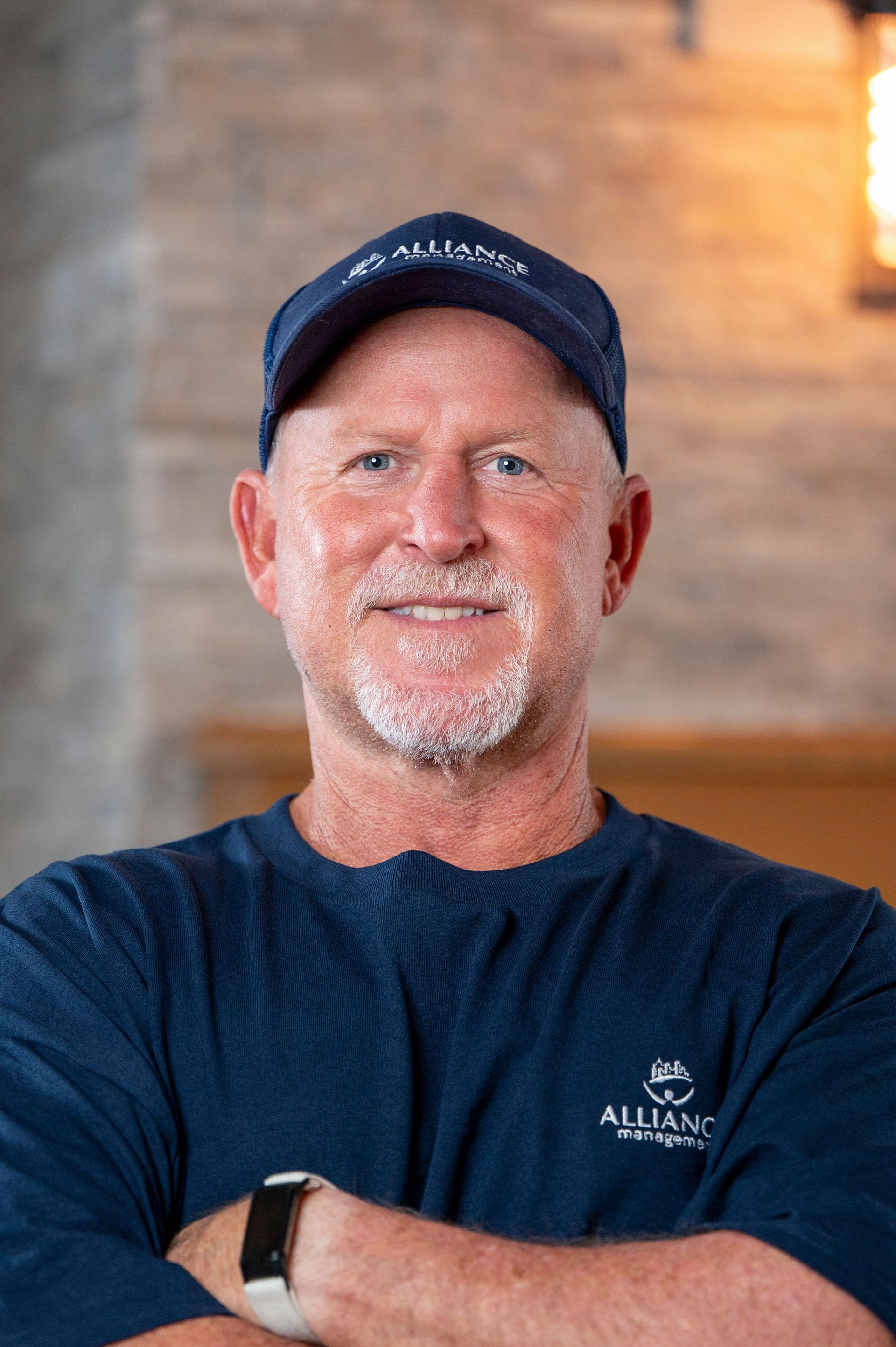 Smiling man in navy cap and shirt with arms crossed, standing indoors against a warm wooden background