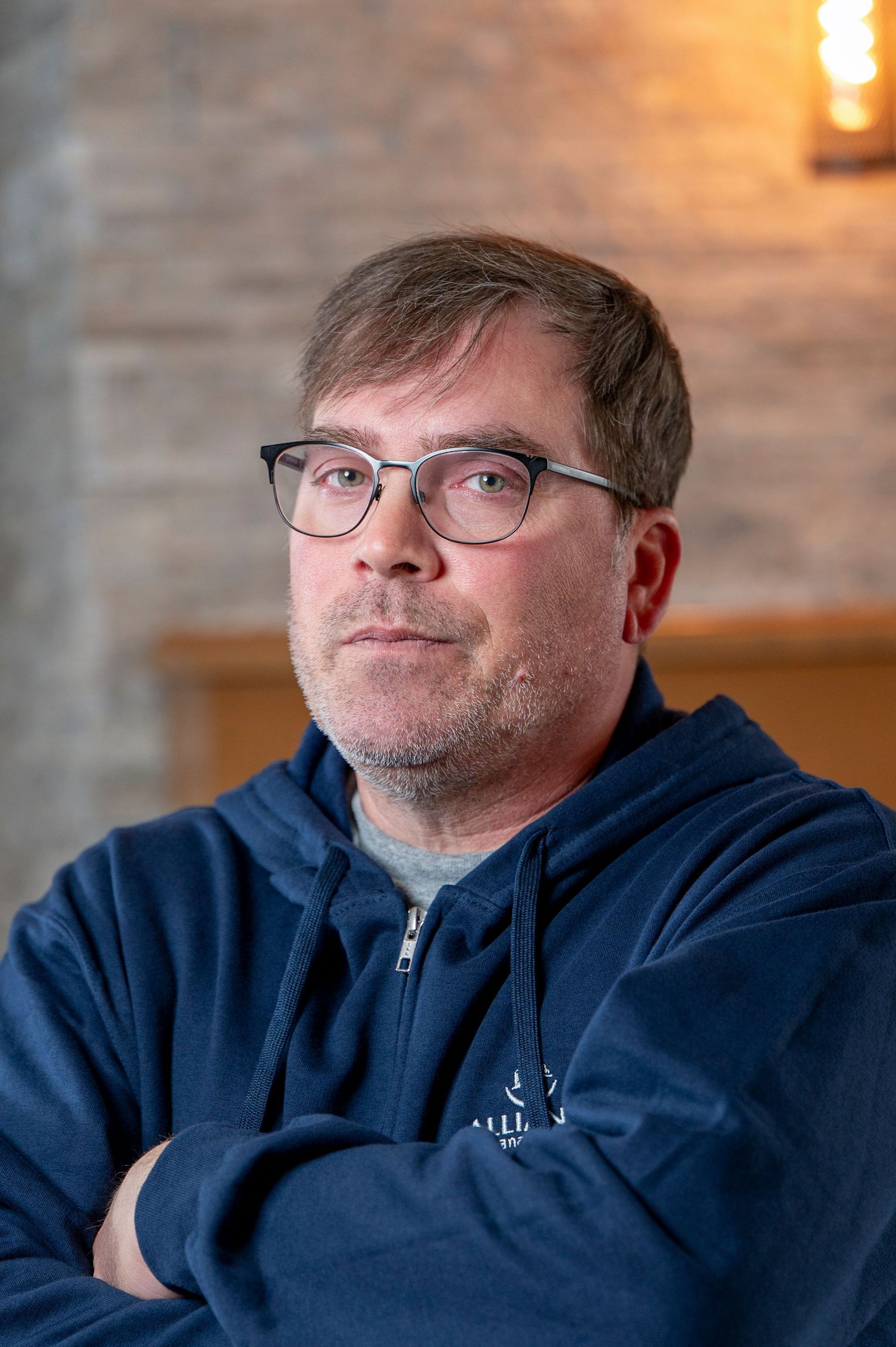 Man with glasses and a blue hoodie, arms crossed, standing indoors against a softly lit background
