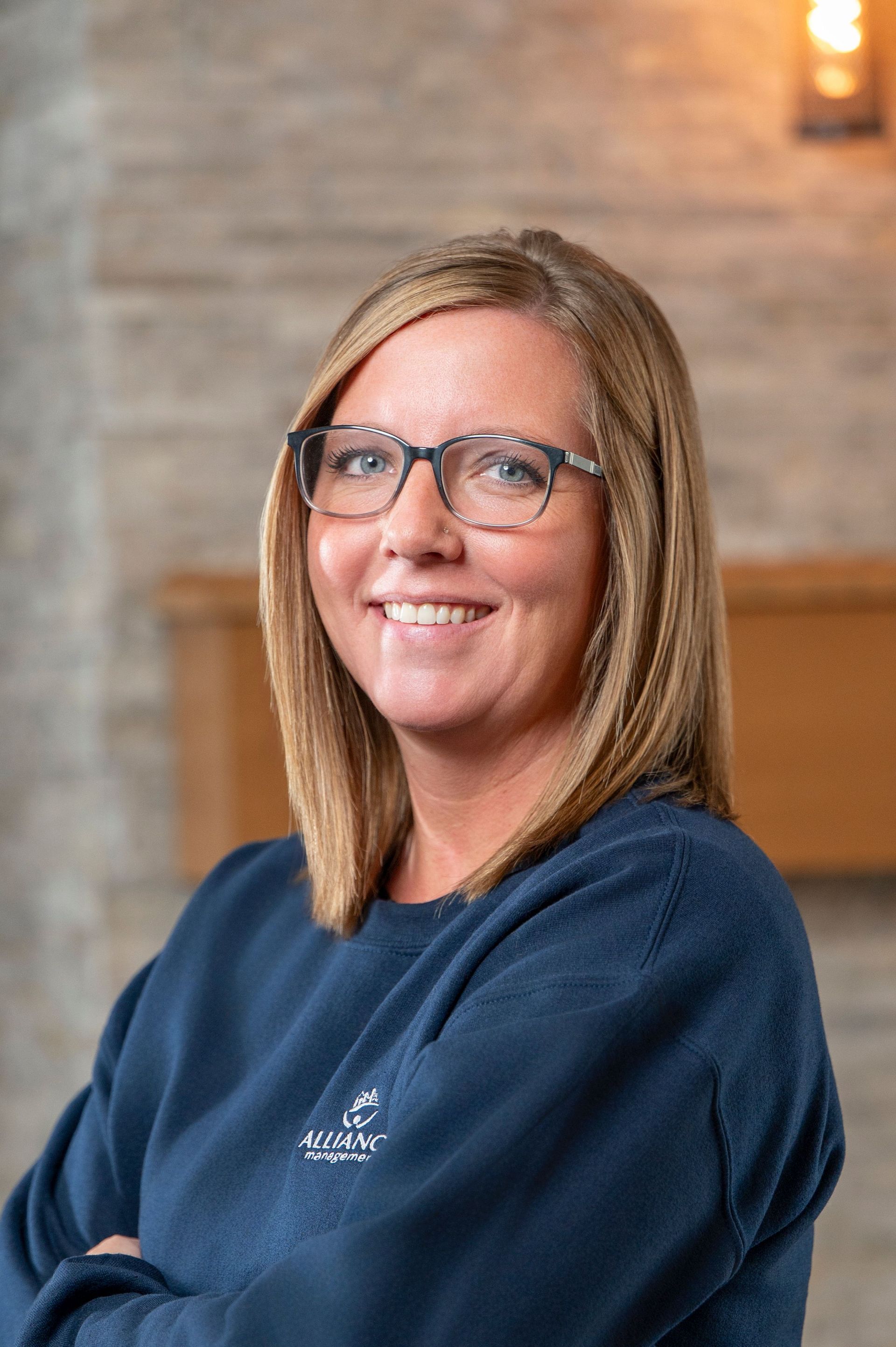 Smiling woman with glasses in a blue sweater, seated indoors against a blurred background