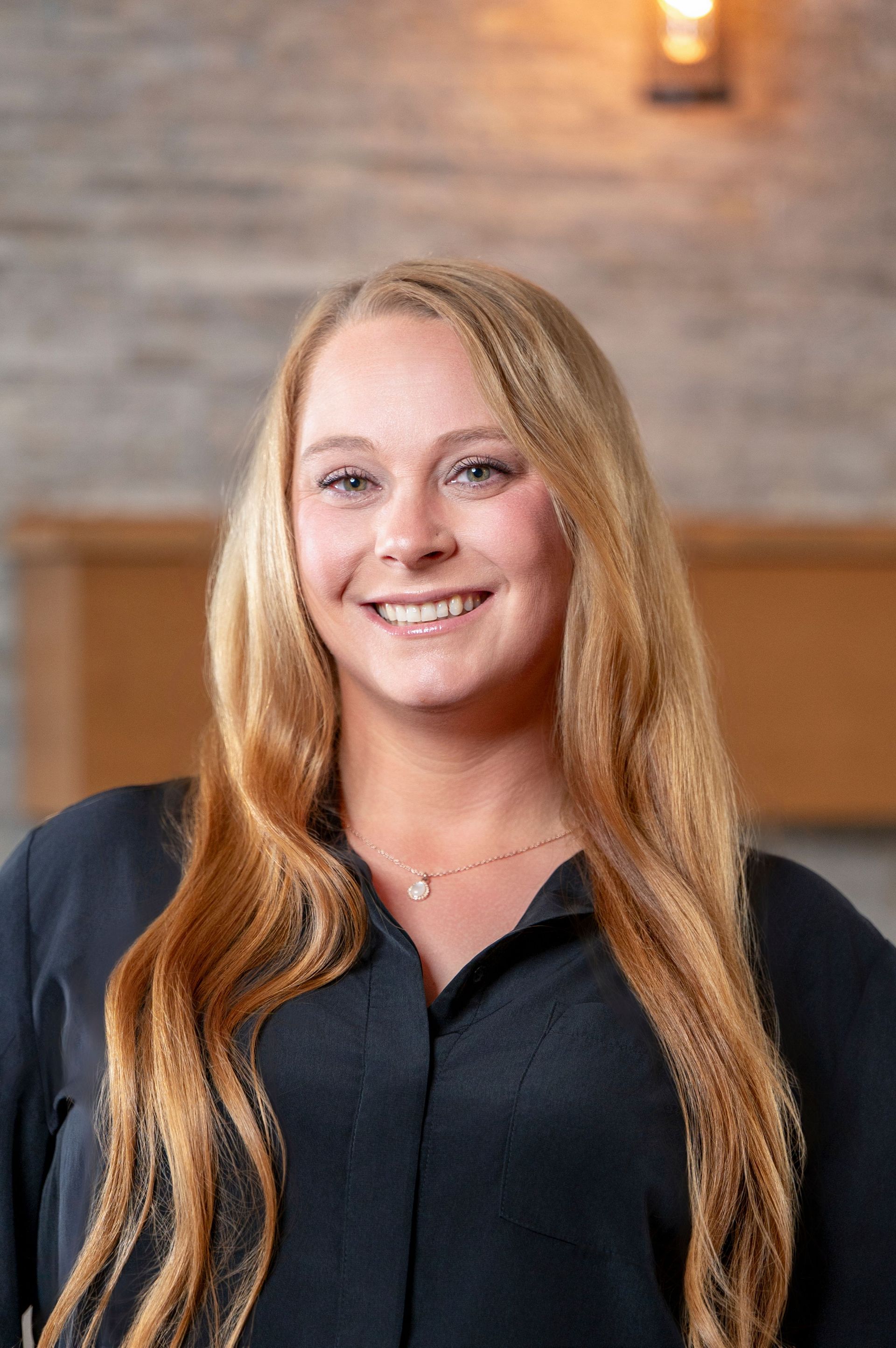 Smiling person with long blonde hair in a dark blouse, standing indoors against a softly lit background